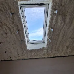 Poddasze z oknem dachowym ocieplone pianką PUR, widoczne metalowe mocowania. Biała piana szczelnie wypełnia przestrzeń wokół ramy okiennej, błękitne niebo w tle.