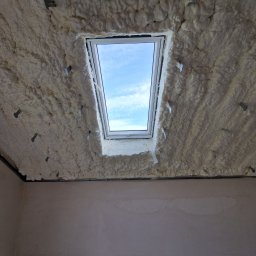 Firma Ogólnobudowlana Michał Łazowski - Poddasze po ociepleniu pianką PUR, widoczne białe, nierówne warstwy piany wokół okna dachowego i metalowe mocowania. W tle ściana przed wykończeniem.