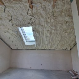 Firma Ogólnobudowlana Michał Łazowski - Poddasze po ociepleniu pianką PUR, widoczne drewniane elementy konstrukcyjne i okno dachowe. Na podłodze leżą kable. Ściany przygotowane do dalszych prac wykończeniowych.