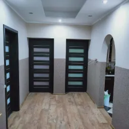 Korytarz po remoncie z trzema ciemnymi drzwiami z przeszkleniami, wykończony drewnopodobną podłogą i strukturalną tapetą w odcieniach szarości, z widocznym fragmentem zabudowy sufitowej...