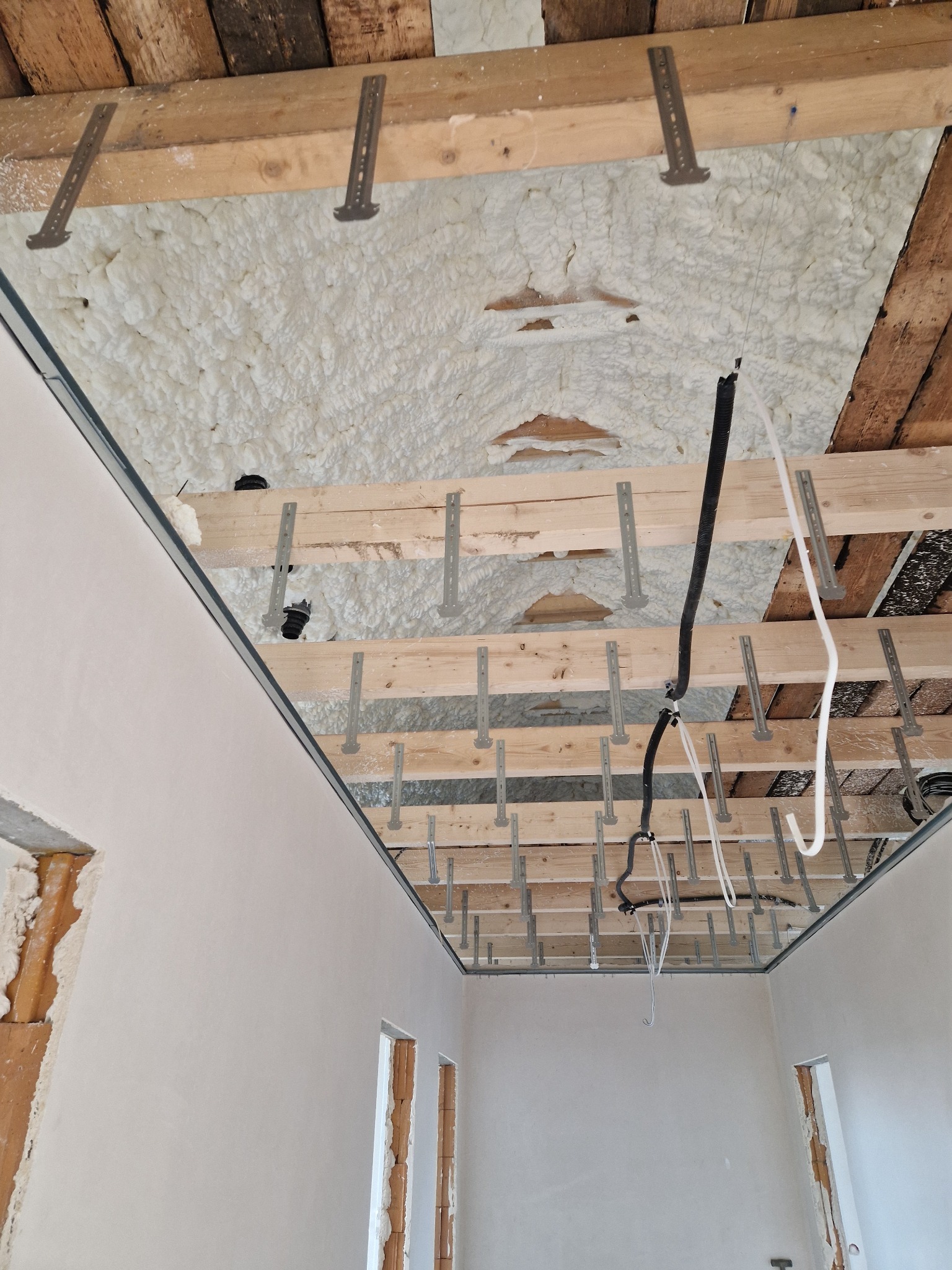 Poddasze ocieplone pianką PUR, widoczne drewniane belki, metalowe uchwyty, instalacja elektryczna. Ściany w trakcie prac wykończeniowych.