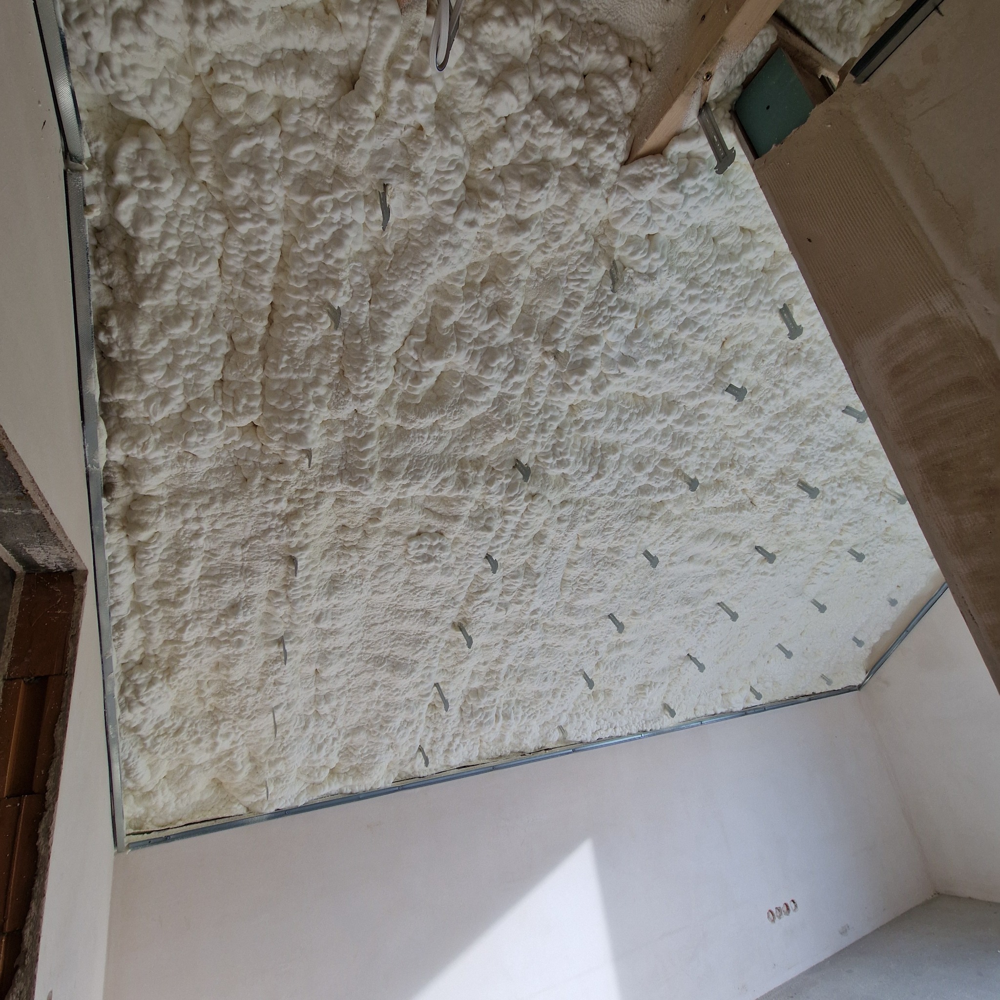 Poddasze po ociepleniu pianką PUR, widoczne metalowe profile mocujące i wystające kołki, jasne wnętrze w trakcie wykańczania.