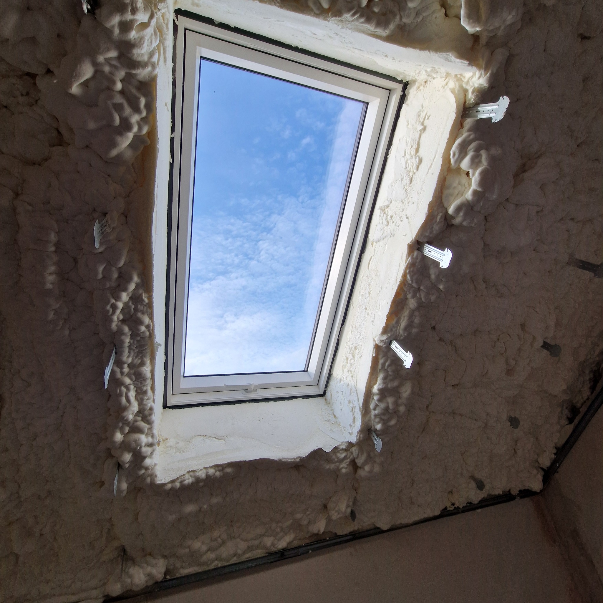 Poddasze z oknem połaciowym po ociepleniu pianką PUR, widoczne metalowe uchwyty mocujące, błękitne niebo przez okno.