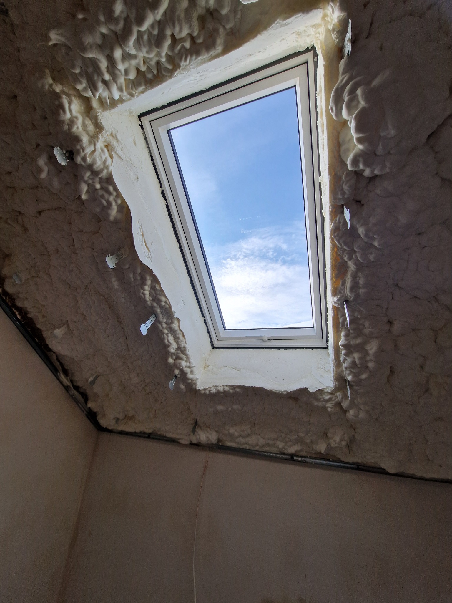 Poddasze z oknem dachowym po ociepleniu pianką PUR, widoczne uchwyty mocujące. Błękitne niebo przez okno. Wykończenie pianką o nieregularnej fakturze.