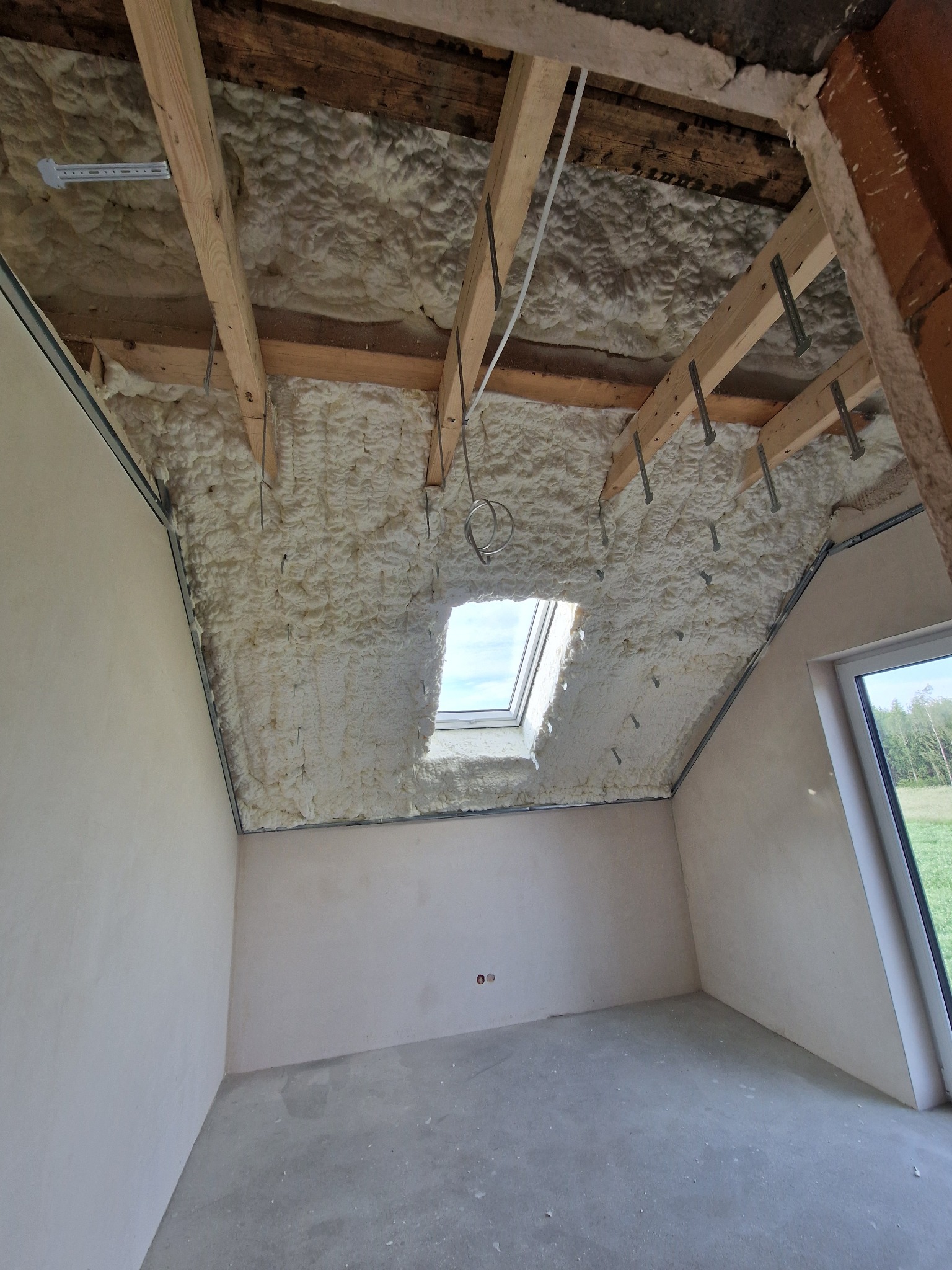 Poddasze po ociepleniu pianką PUR, widoczne drewniane elementy konstrukcyjne i okno dachowe. Biała pianka szczelnie wypełnia przestrzeń między krokwiami.