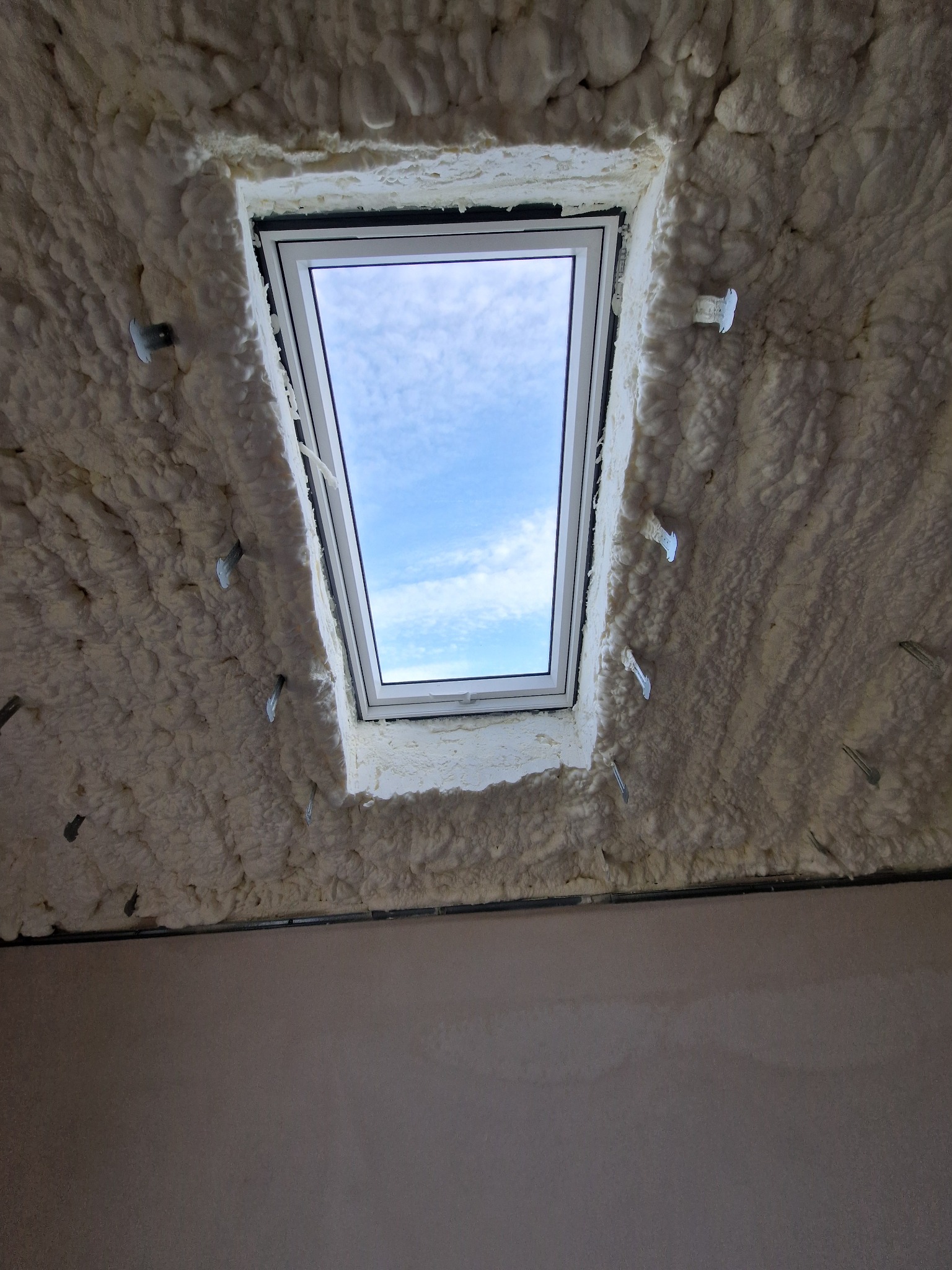 Poddasze z oknem dachowym ocieplone pianką PUR, widoczne metalowe mocowania. Biała piana szczelnie wypełnia przestrzeń wokół ramy okiennej, błękitne niebo w tle.