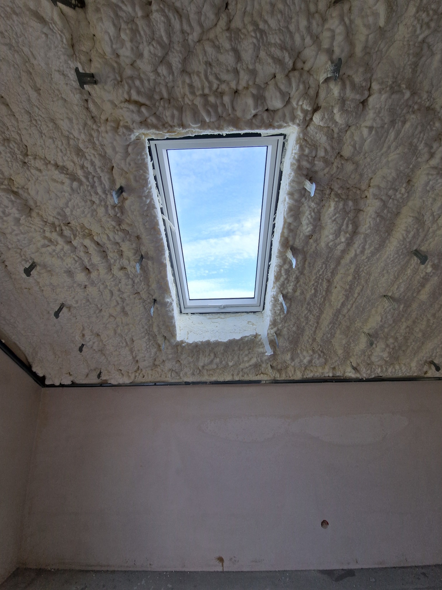Poddasze po ociepleniu pianką PUR, widoczne białe, nierówne warstwy piany wokół okna dachowego i metalowe mocowania. W tle ściana przed wykończeniem.