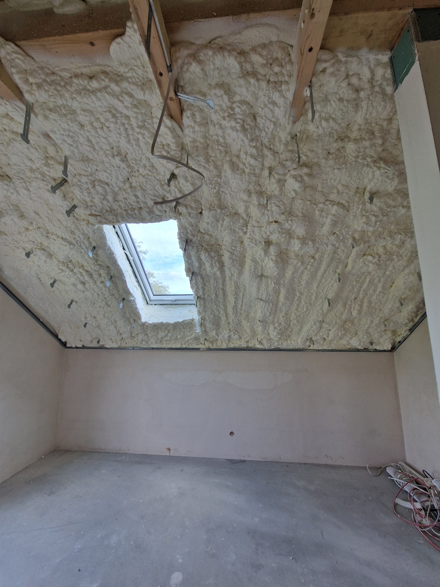 Poddasze po ociepleniu pianką PUR, widoczne drewniane elementy konstrukcyjne i okno dachowe. Na podłodze leżą kable. Ściany przygotowane do dalszych prac wykończeniowych.