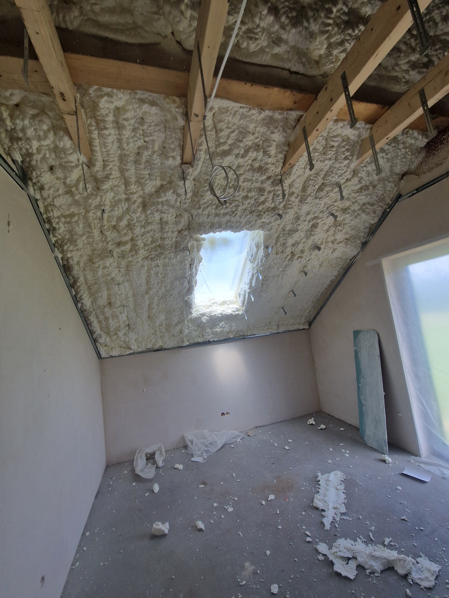 Poddasze po ociepleniu pianką PUR, widoczne drewniane krokwie i okno dachowe. Na podłodze resztki piany, ściany przygotowane do dalszych prac wykończeniowych.