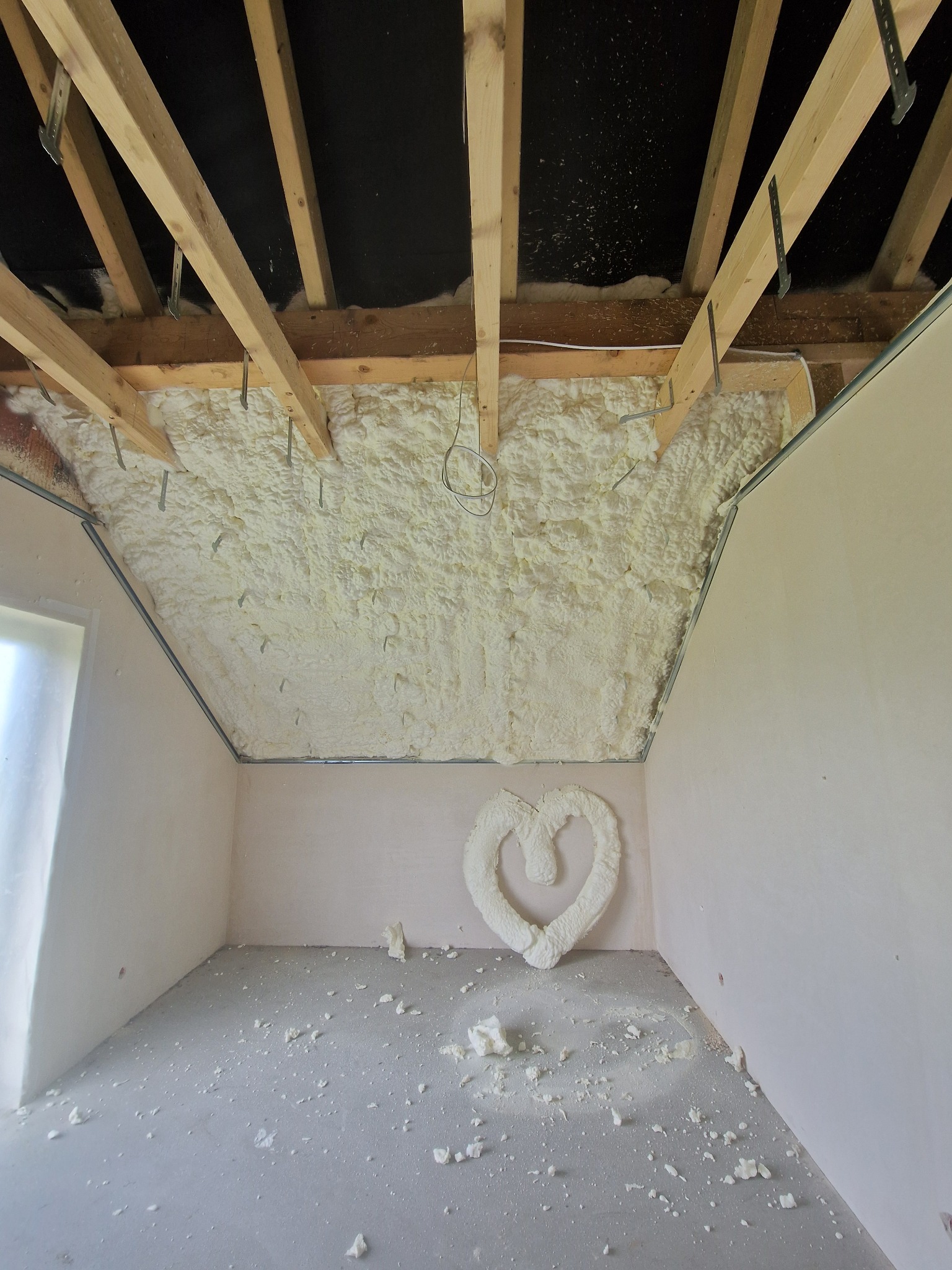 Poddasze po ociepleniu pianką PUR; widoczne drewniane krokwie, instalacja elektryczna i dekoracja w kształcie serca z pianki. Fragmenty pianki na podłodze.