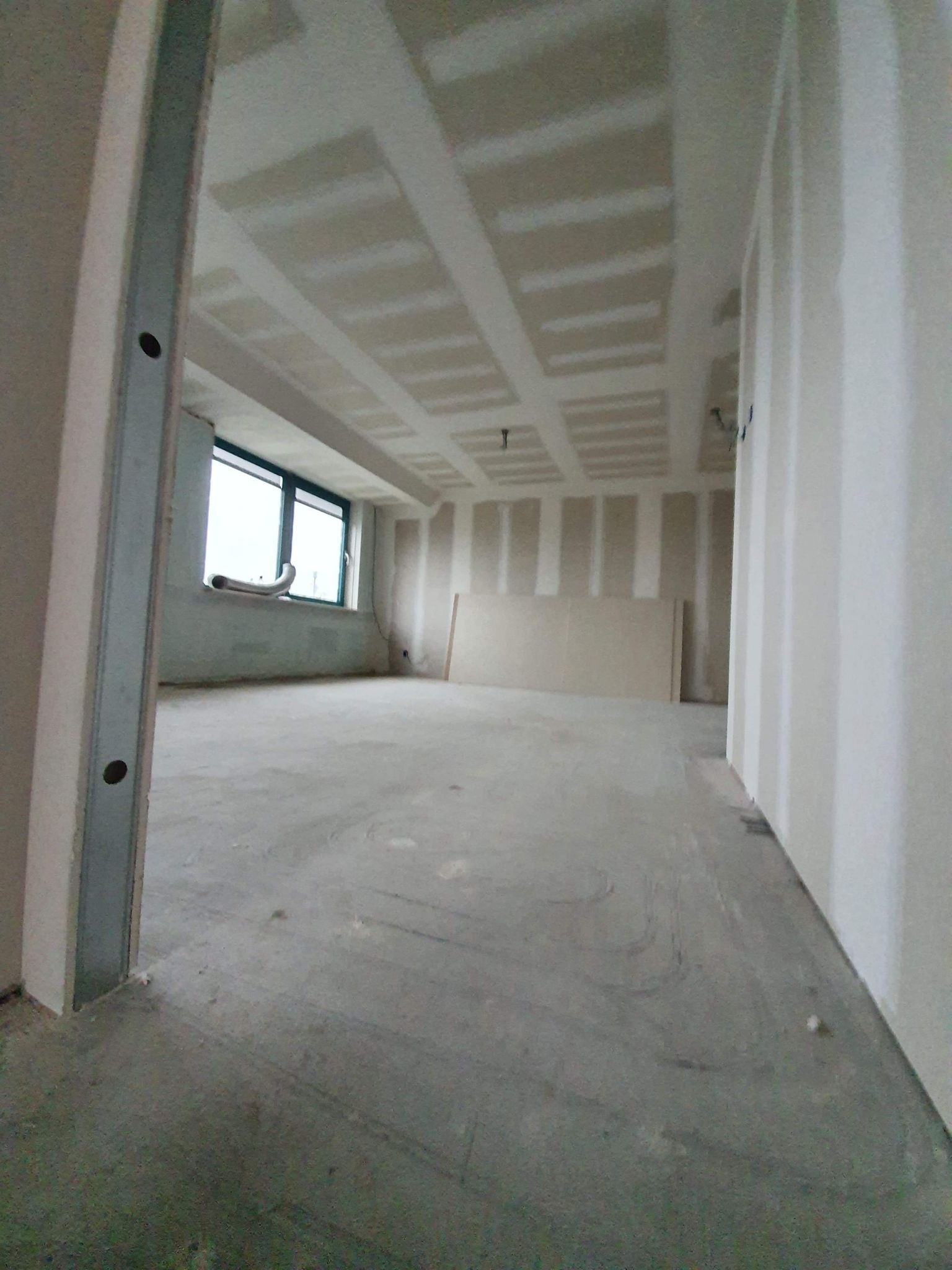 Wnętrze w trakcie wykańczania: widoczne metalowe profile, płyty kartonowo-gipsowe na ścianach i suficie, surowy beton na podłodze, okno z wystającym elementem instalacji wentylacyjnej.