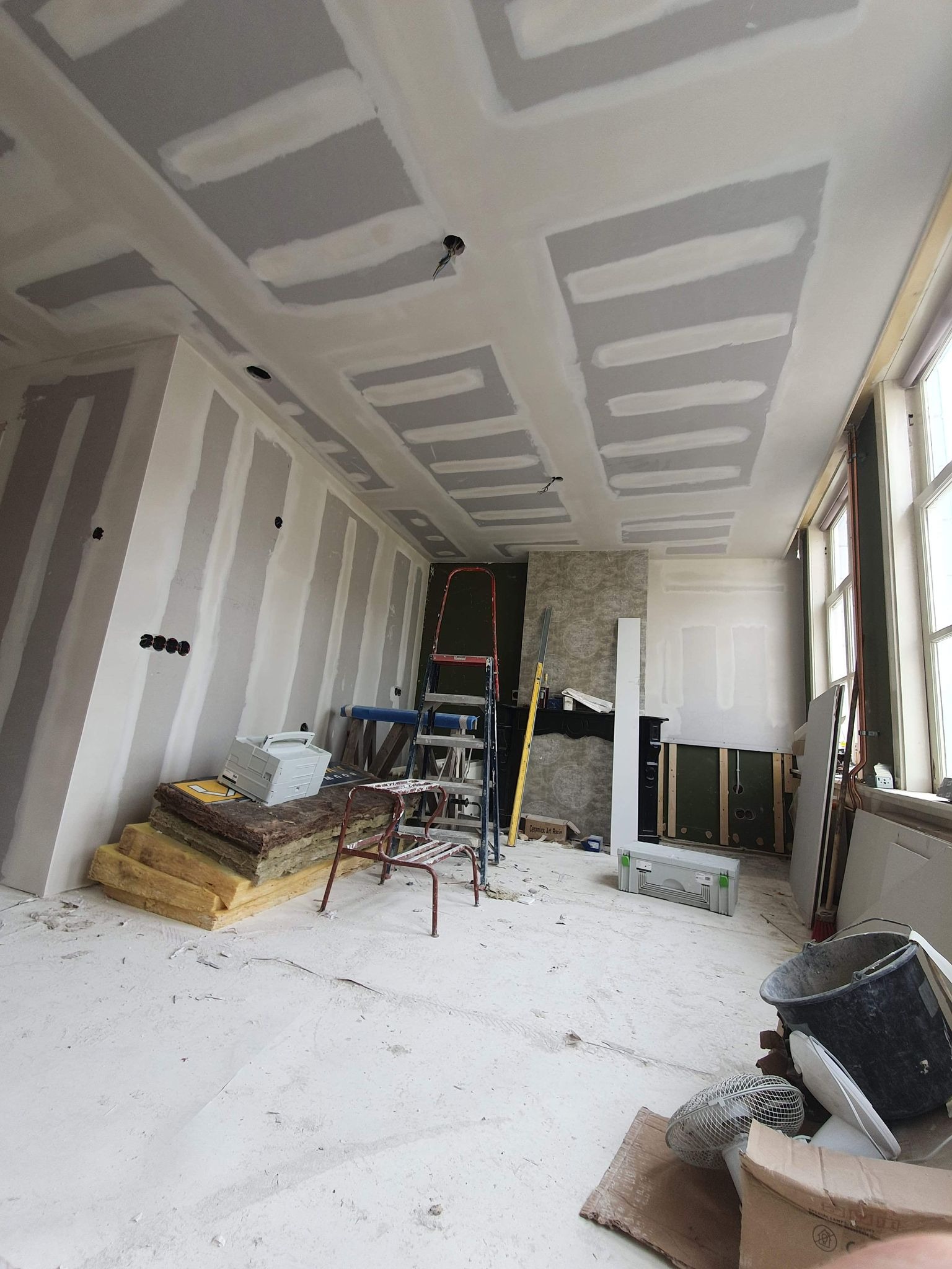 Pokój w trakcie remontu z widocznymi płytami kartonowo-gipsowymi na ścianach i suficie, częściowo wyszpachlowanymi, z rozstawioną drabiną i materiałami budowlanymi na podłodze.