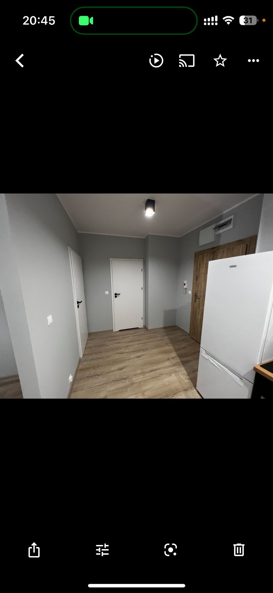 Szare ściany w odnowionym pokoju z drewnopodobną podłogą, białe drzwi z czarną klamką i drewniane drzwi obok białej lodówki.