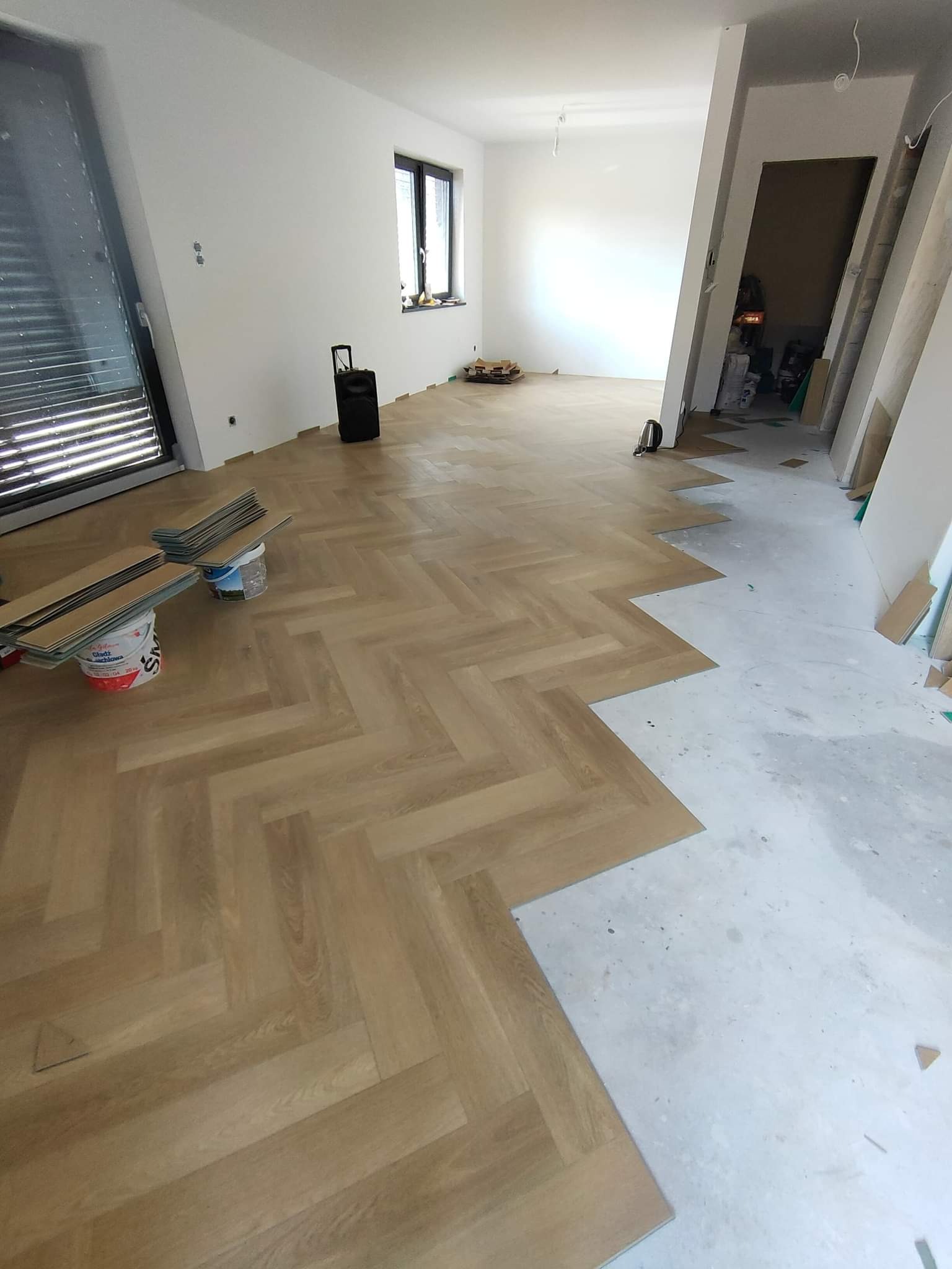 Układanie parkietu w jodełkę w jasnym, przestronnym wnętrzu. Część podłogi już wykończona, widoczne panele i wiaderko z klejem.