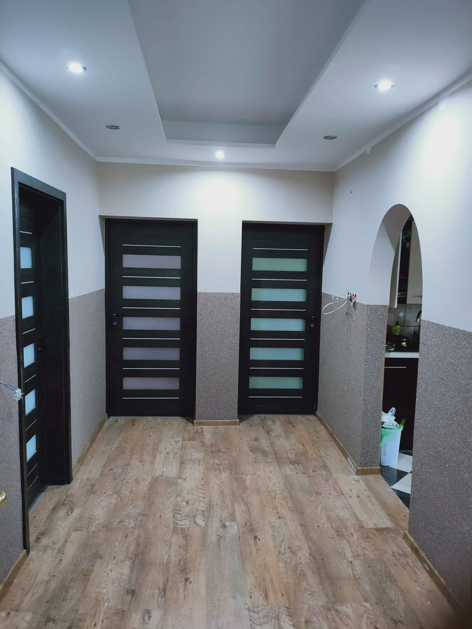 Korytarz po remoncie z trzema ciemnymi drzwiami z przeszkleniami, wykończony drewnopodobną podłogą i strukturalną tapetą w odcieniach szarości, z widocznym fragmentem zabudowy sufitowej...