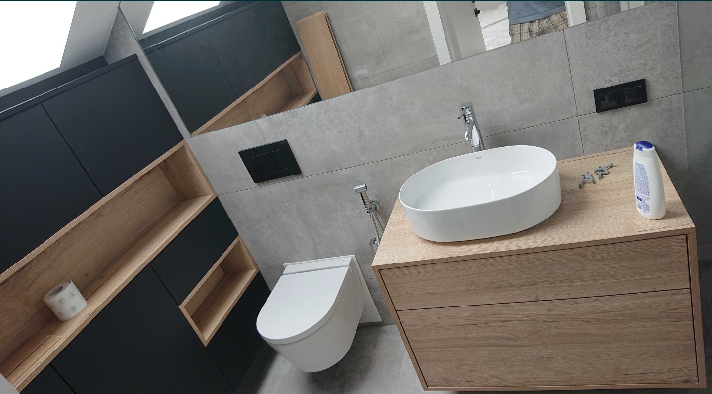 Nowoczesna łazienka z wiszącą toaletą, owalną umywalką na drewnianej szafce, szarą płytką ścienną i czarnymi szafkami z drewnianymi wnękami. Na blacie leży butelka płynu i elementy montażowe.