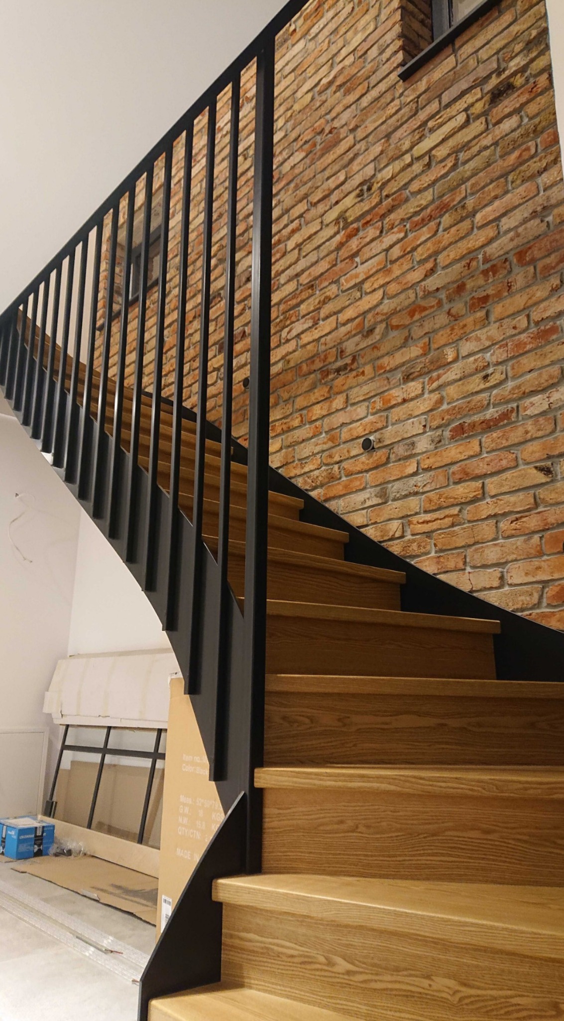 Drewniane schody z czarną metalową balustradą na tle ściany z cegły, widoczne kartony i materiały budowlane w tle.