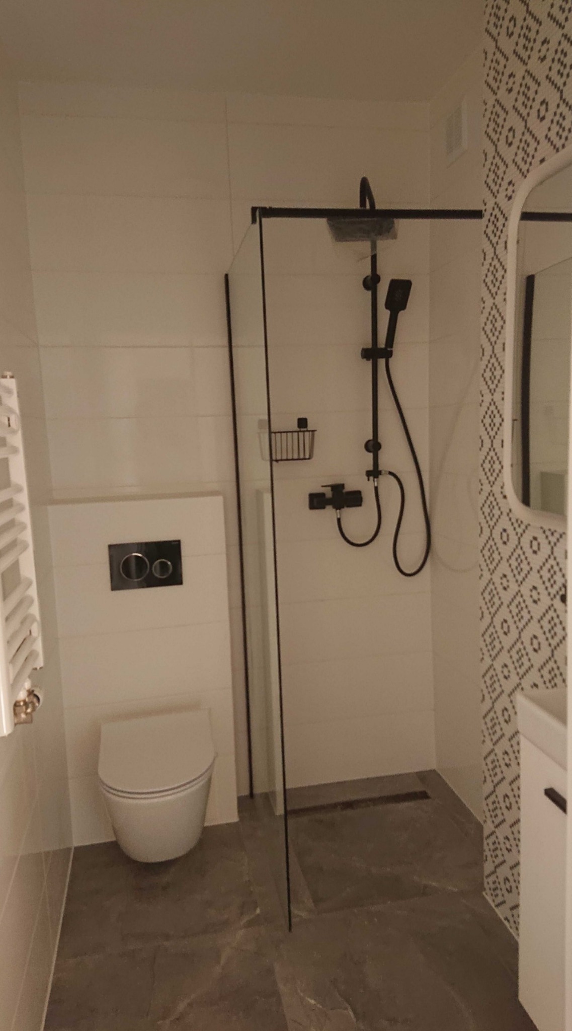 Nowoczesna łazienka z kabiną prysznicową typu walk-in z czarną armaturą, podwieszana toaleta z czarnym przyciskiem, grzejnik drabinkowy i szare płytki imitujące kamień na podłodze.