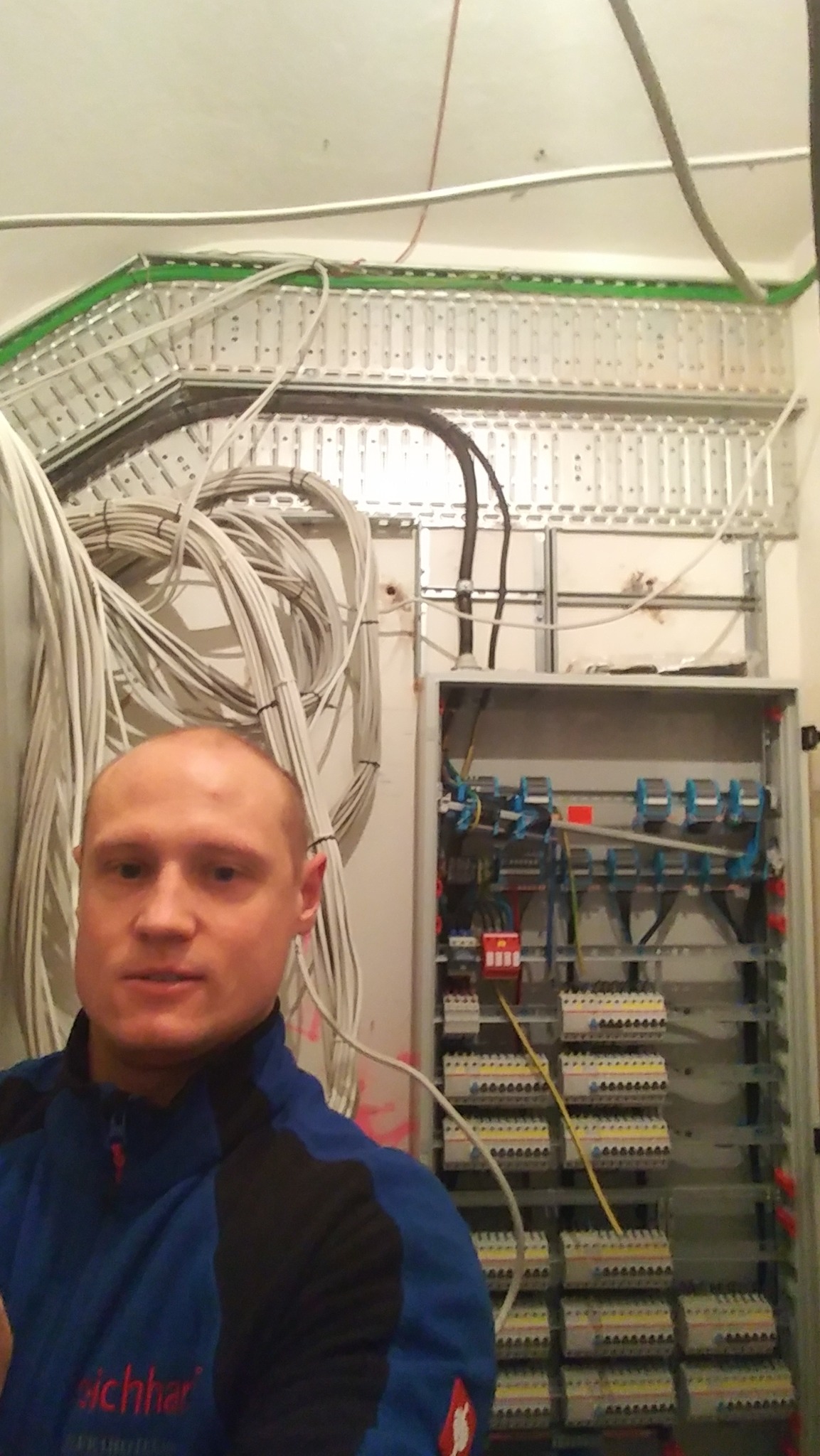 Elektryk prezentuje rozdzielnicę elektryczną z licznymi zabezpieczeniami i okablowaniem w korytach metalowych.