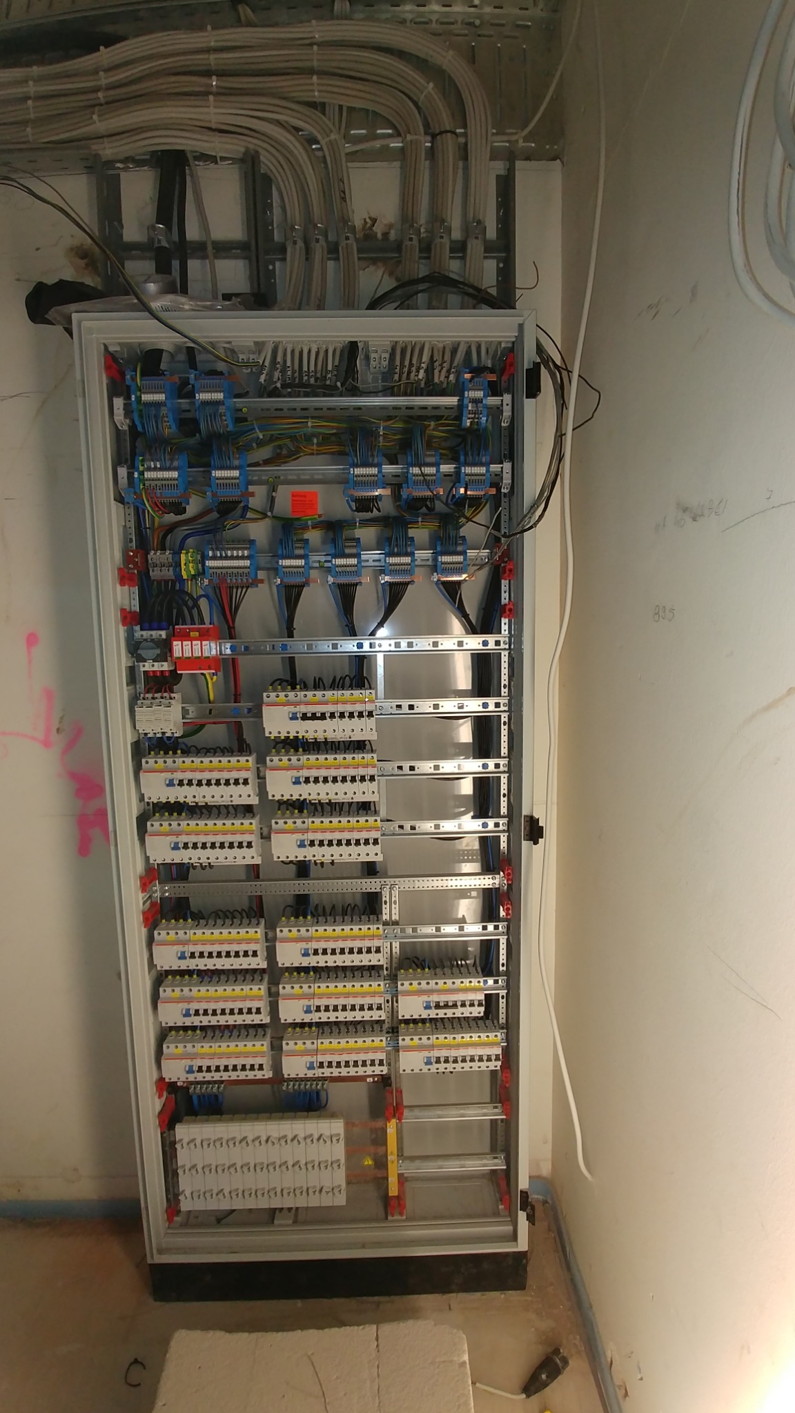 Otwarta szafa elektryczna z rozdzielnicą, widoczne liczne przewody, bezpieczniki, listwy zaciskowe i elementy montażowe, z przewodem ochronnym u góry