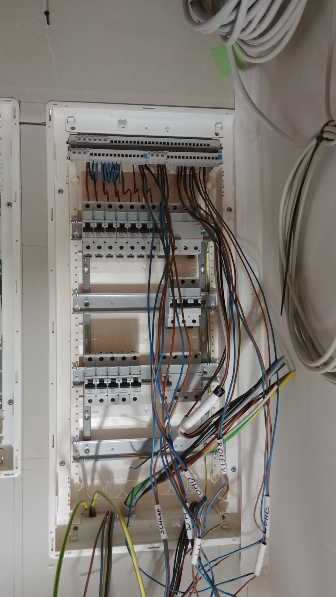 Rozdzielnica elektryczna w trakcie montażu, widoczne liczne przewody z opisami oraz elementy zabezpieczeń, częściowo zamontowane w białej obudowie, z widocznymi elementami montażowymi.