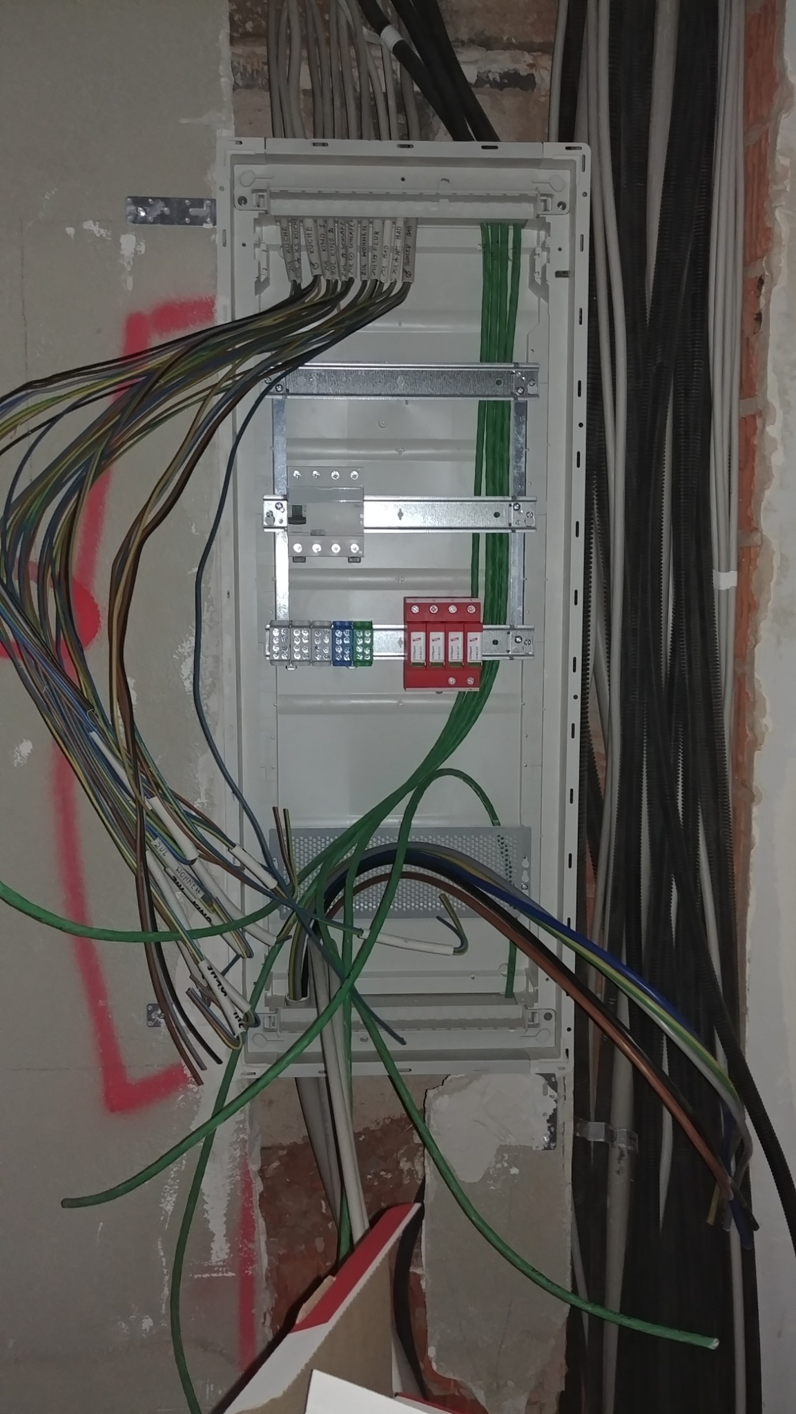 Rozdzielnica elektryczna w trakcie montażu, widoczne kable z oznaczeniami pomieszczeń, szyna DIN z zabezpieczeniami, ściana w stanie surowym z widocznymi śladami po instalacji.