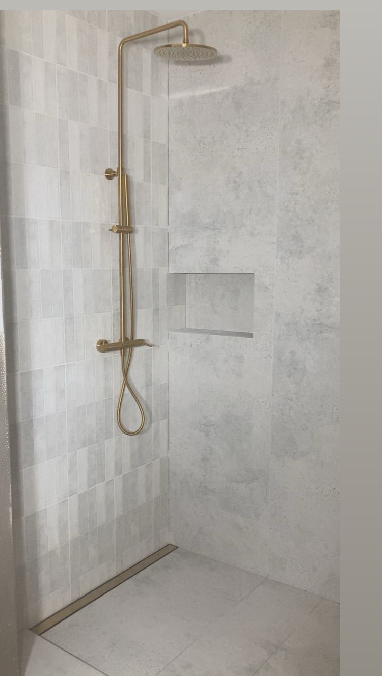 Wykończona kabina prysznicowa z szarymi płytkami w dwóch wzorach, złotą armaturą i odpływem liniowym. W ścianie wnęka. Minimalistyczny design.