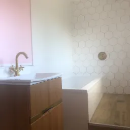 Nowoczesna łazienka z drewnianą szafką pod umywalką z złotą armaturą, białą wanną obłożoną białymi heksagonalnymi płytkami i drewnopodobną podłogą.