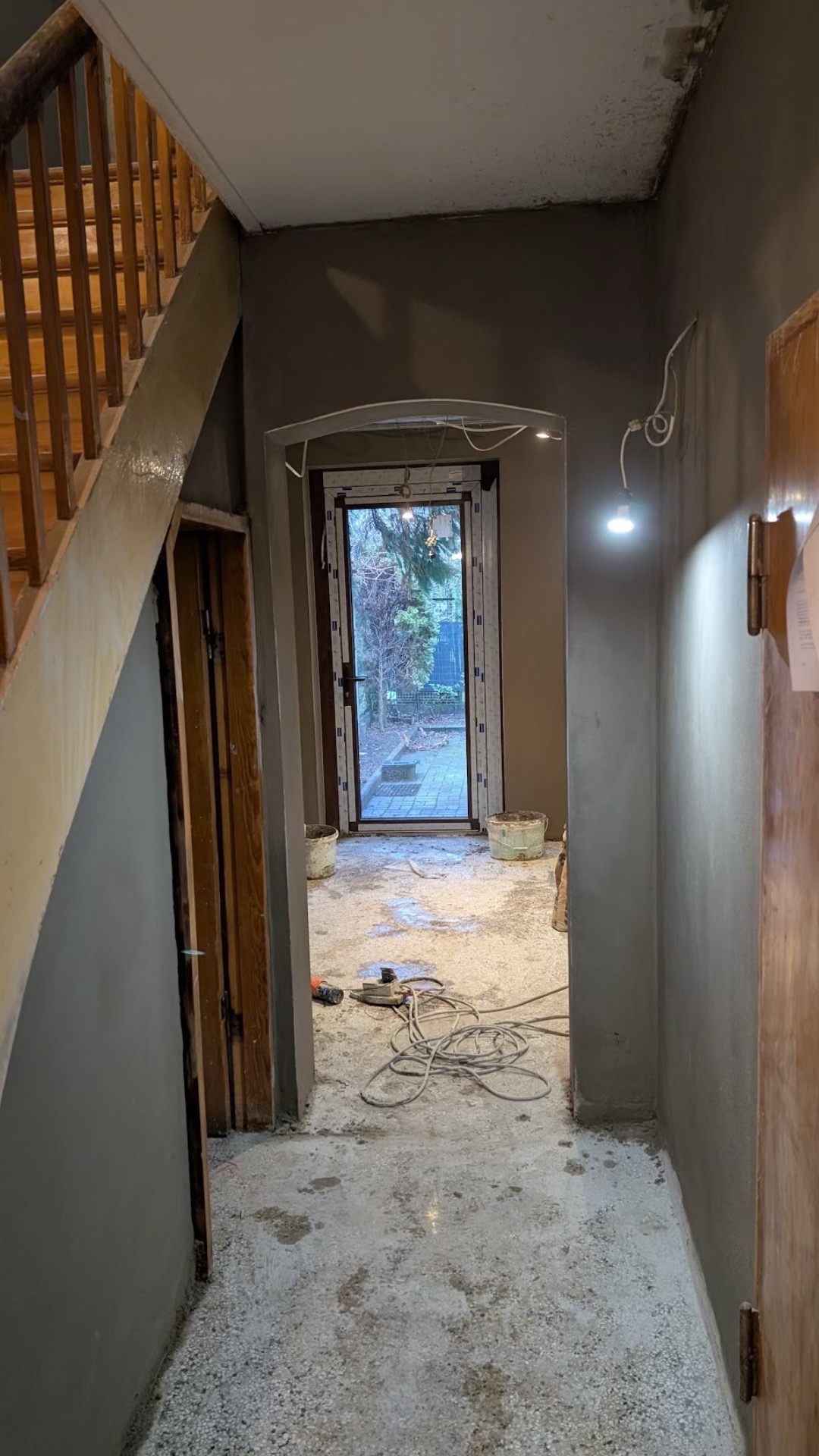 Wnętrze korytarza w trakcie prac remontowych: świeżo otynkowane ściany w odcieniach szarości, widoczne narzędzia i kable na podłodze, fragment drewnianych schodów po lewej stronie oraz drzwi...