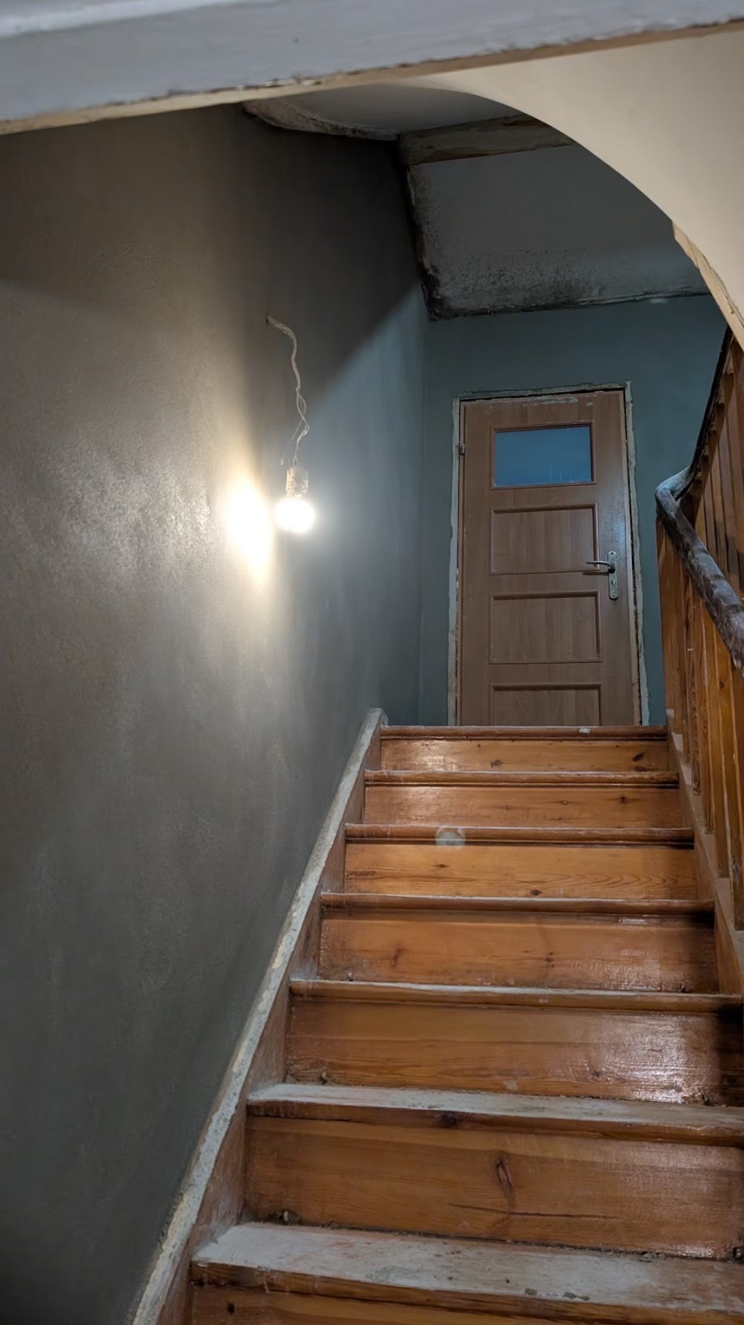 Klatka schodowa w trakcie prac tynkarskich: świeżo otynkowana szara ściana z widocznym połyskiem, drewniane schody zabezpieczone przed zabrudzeniem, surowa żarówka na kablu, drewniane drzwi...