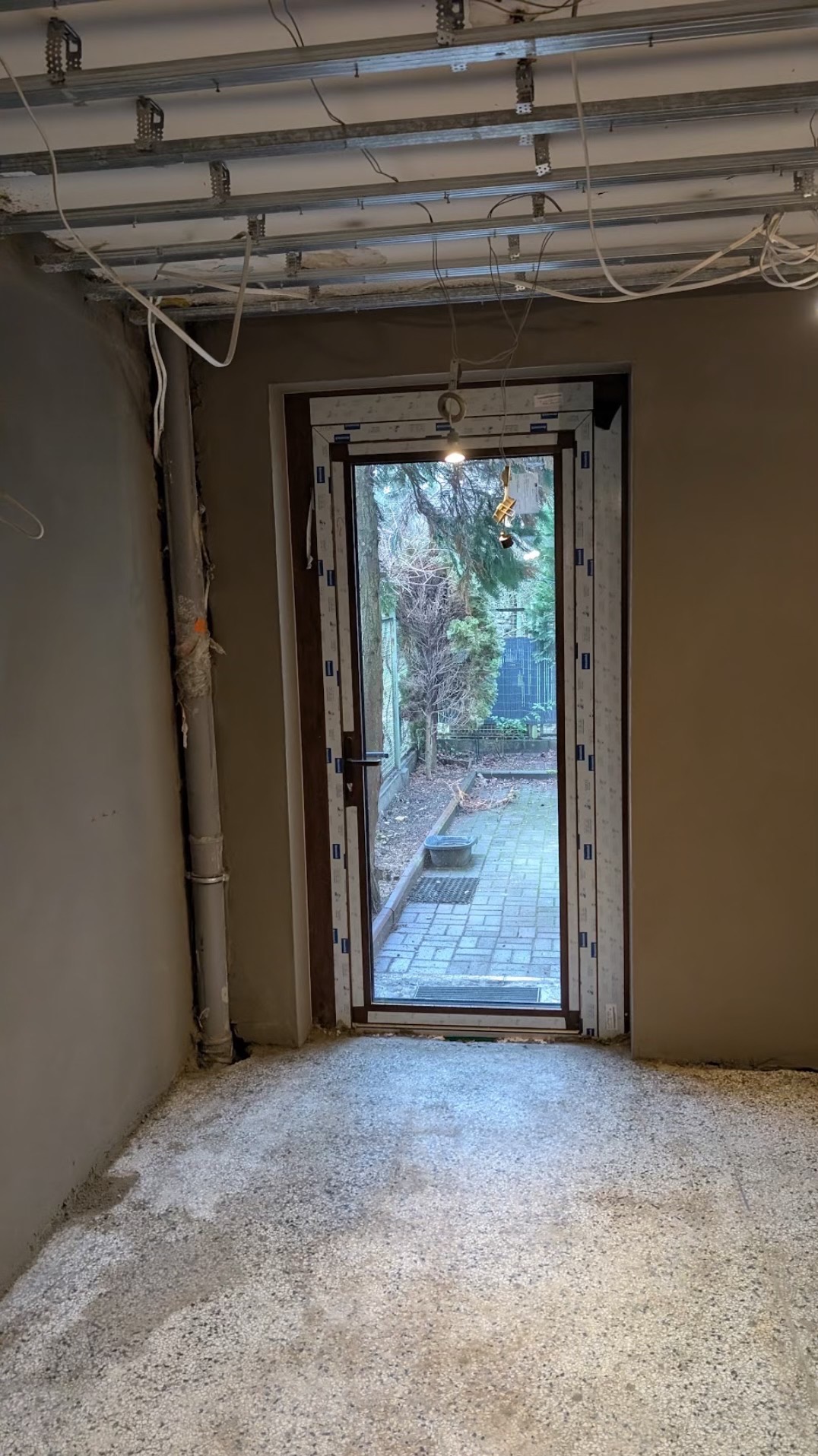 Wnętrze w trakcie prac wykończeniowych: surowe ściany z widocznymi przewodami, wylewka na podłodze, widok na ogród przez otwarte drzwi wejściowe.