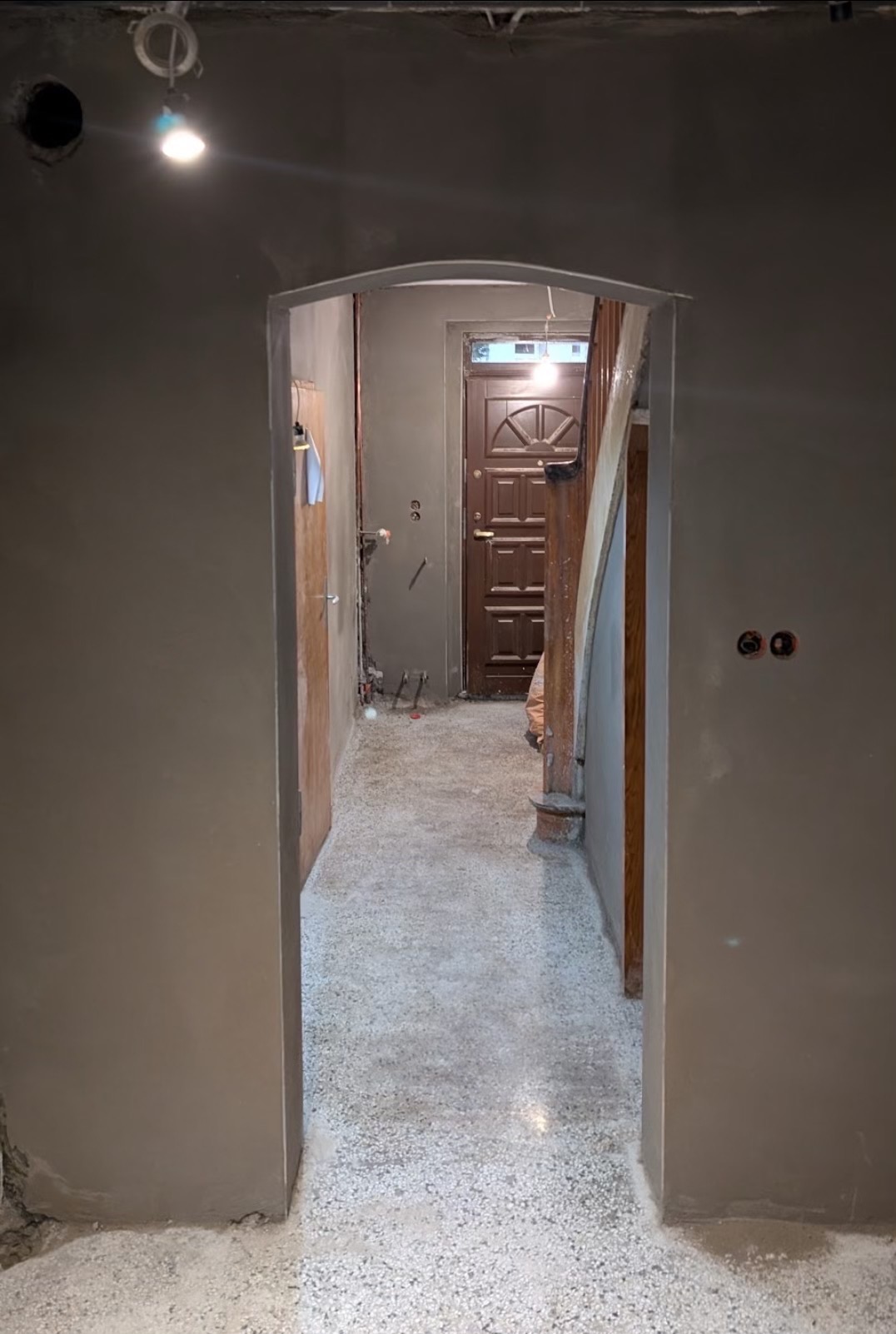 Korytarz w trakcie prac wykończeniowych: ściany pokryte świeżym, szarym tynkiem, widoczne instalacje elektryczne i rury, surowy beton na podłodze, drzwi wejściowe w tle.