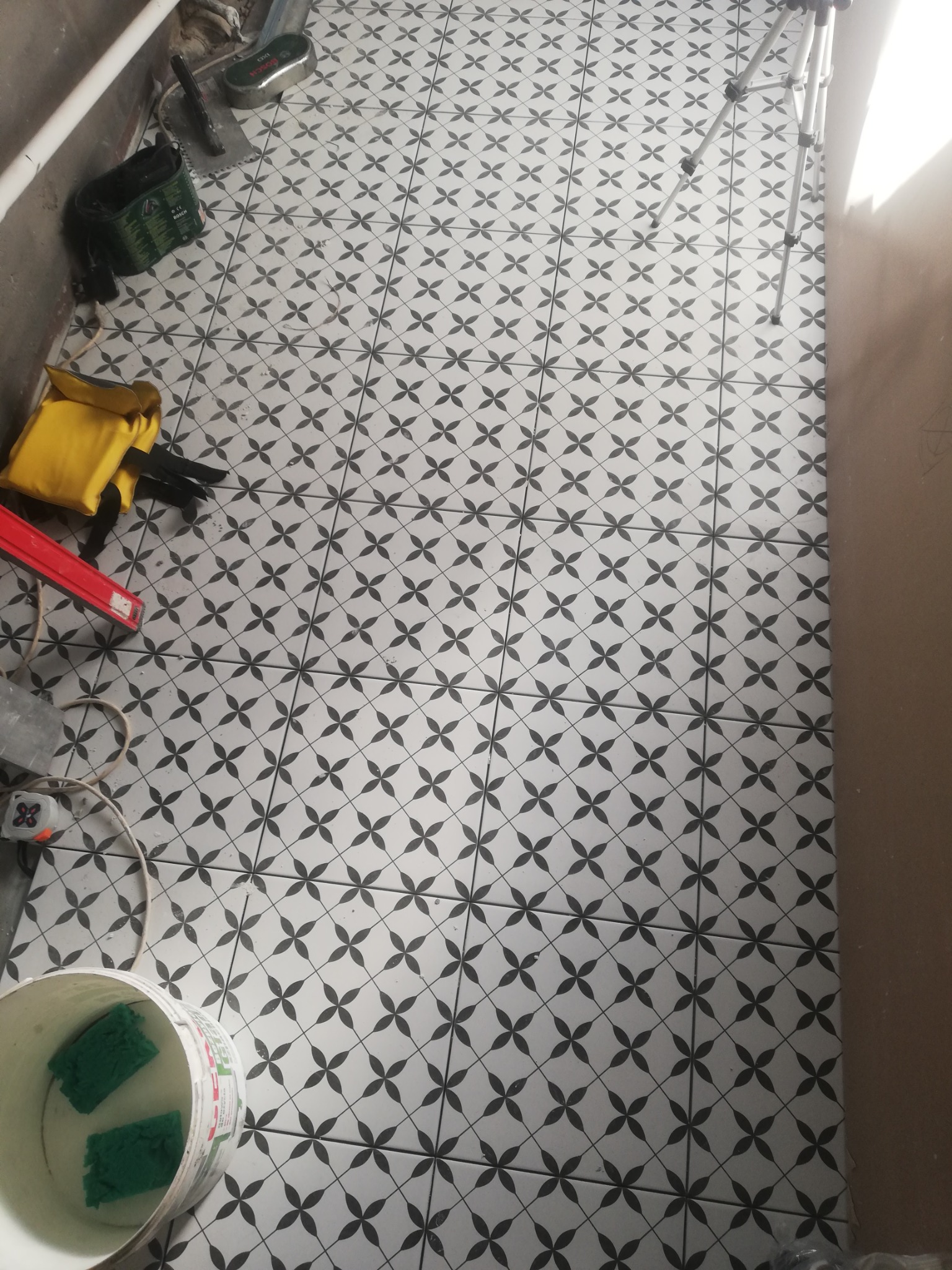 Ułożona podłoga z płytek w czarno-biały wzór kwiatowy, widoczne narzędzia glazurnicze: poziomica, wiadro z gąbkami, szpachelka i torba narzędziowa, w tle fragment ściany i statyw.