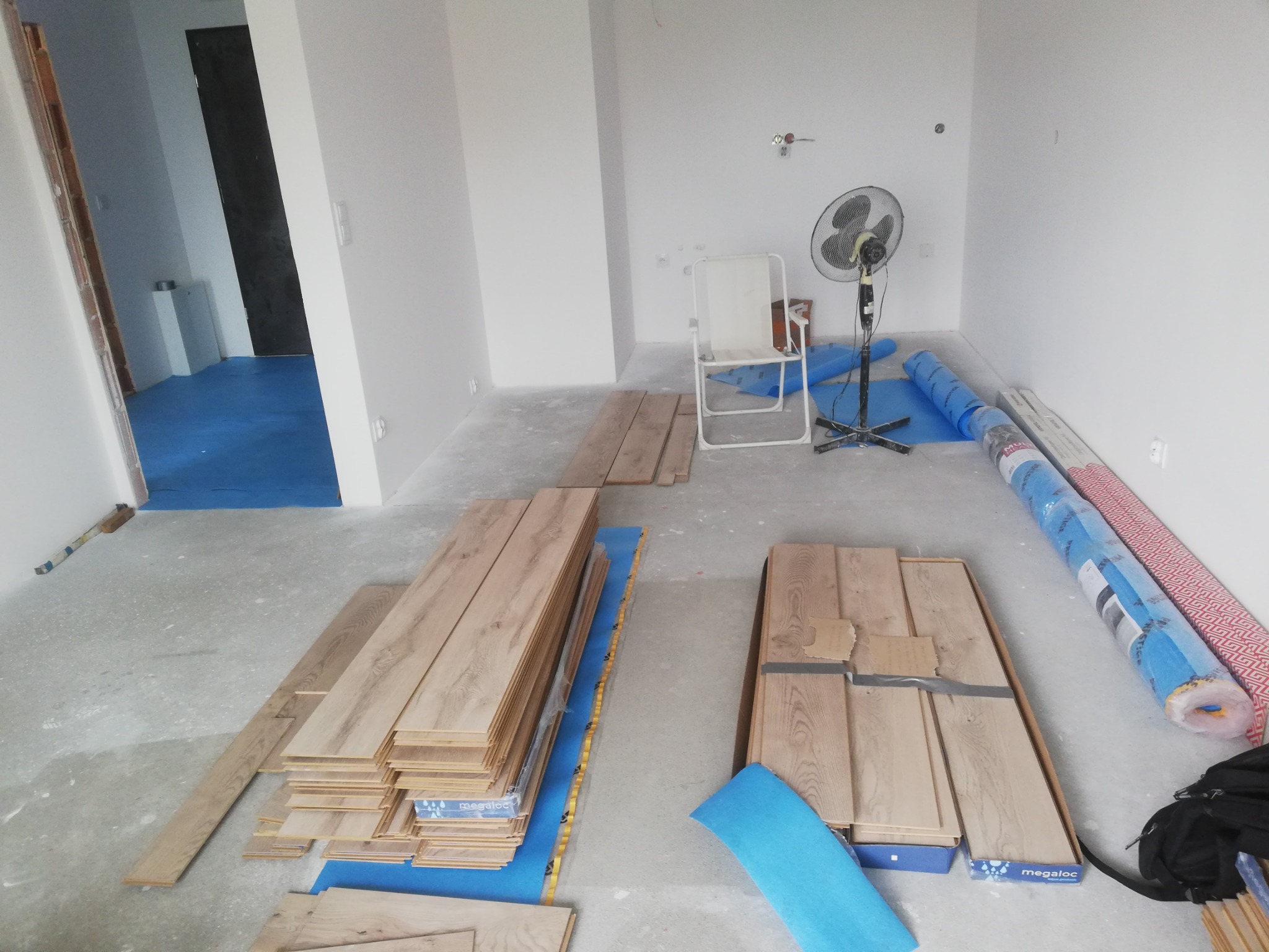 Przygotowanie do montażu paneli podłogowych: deski ułożone na niebieskiej folii, w tle białe ściany, krzesło turystyczne i wentylator stojący.