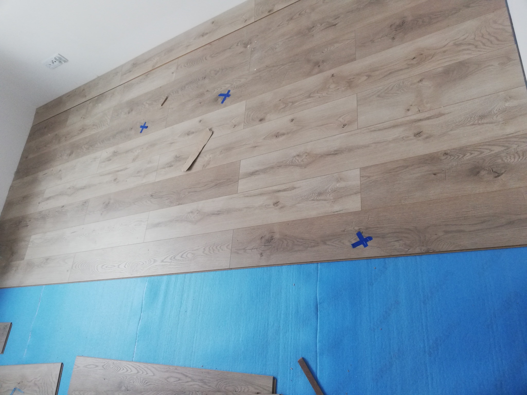 Montaż paneli ściennych z imitacją drewna, zabezpieczonych niebieską folią ochronną, fragment oderwanego kartonu i niebieska taśma maskująca.