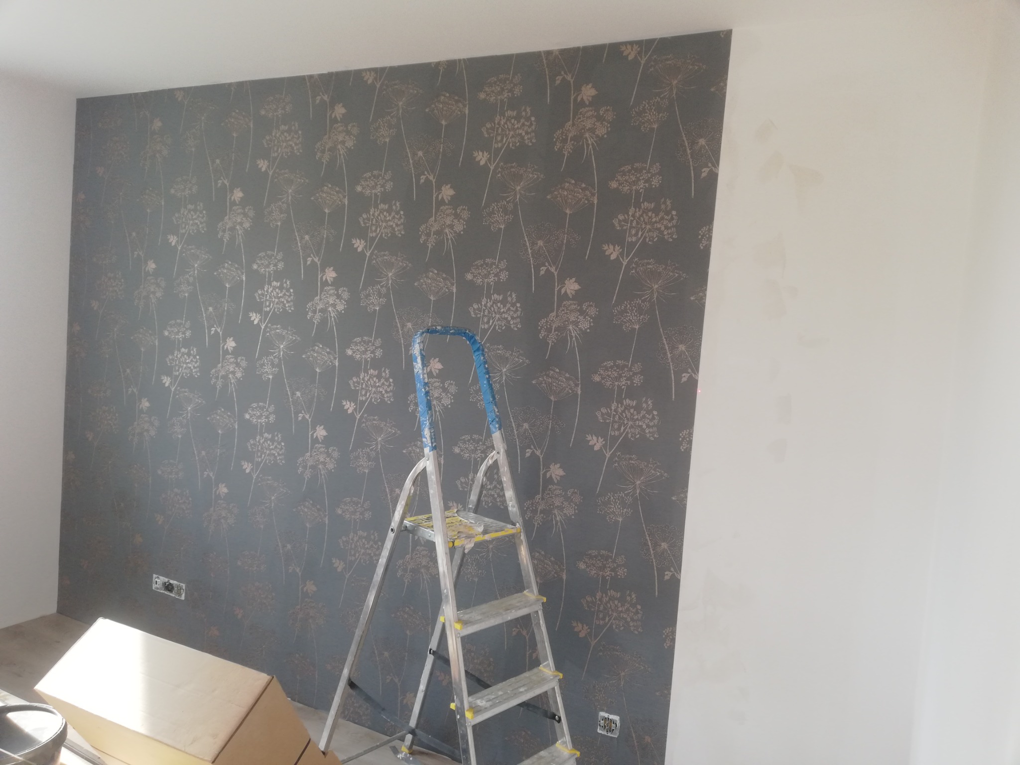 Świeżo położona tapeta w kwiatowy wzór na szarej ścianie, widoczna aluminiowa drabina i fragment białej ściany w trakcie przygotowania.