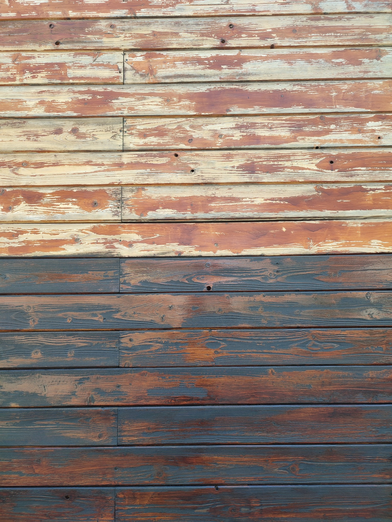 Drewniana ściana z poziomych desek, górna część z resztkami jasnej farby, dolna część pomalowana ciemnobrązową bejcą, widoczne sęki i ślady po gwoździach.