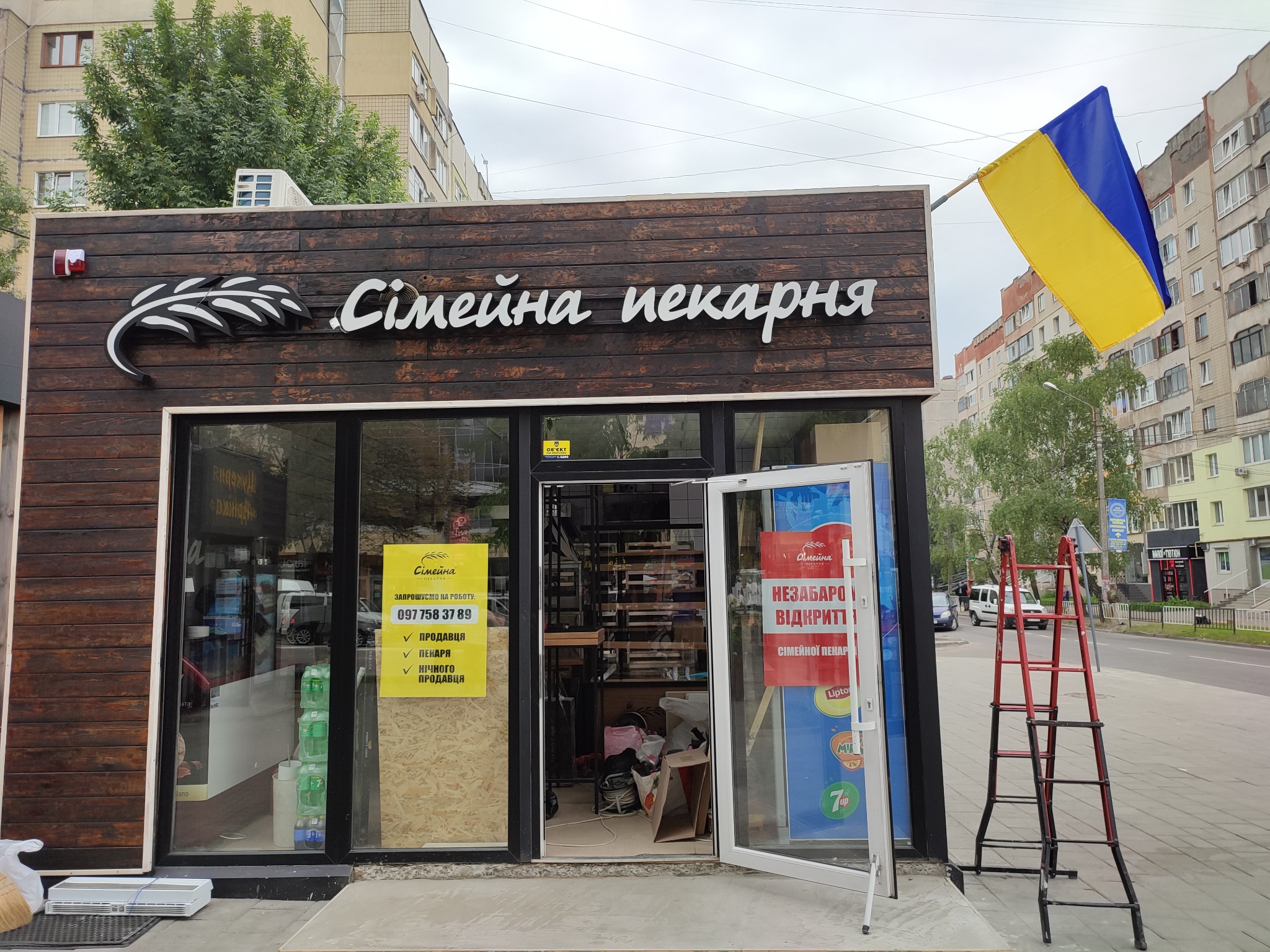 Fasada piekarni z drewnianą okładziną, logo z motywem kłosa i napisem 'Сімейна пекарня', flaga Ukrainy powiewająca nad wejściem, czerwona drabina oparta o ścianę, widoczne ogłoszenia o rekrutacji.