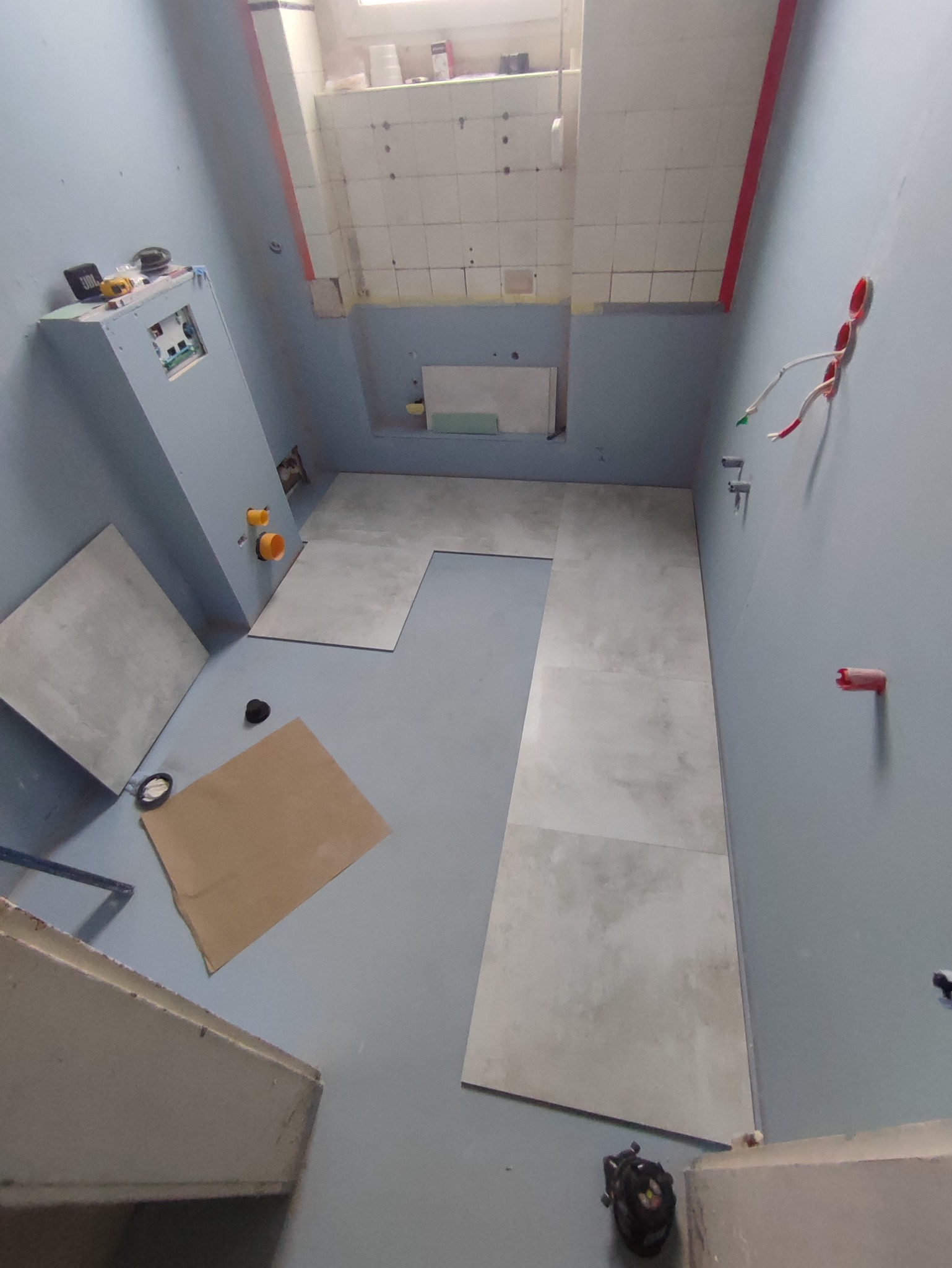 Układanie szarych, matowych płytek podłogowych w surowej łazience w trakcie remontu; widoczne wystające przewody elektryczne, nieotynkowane ściany i narzędzia glazurnicze.