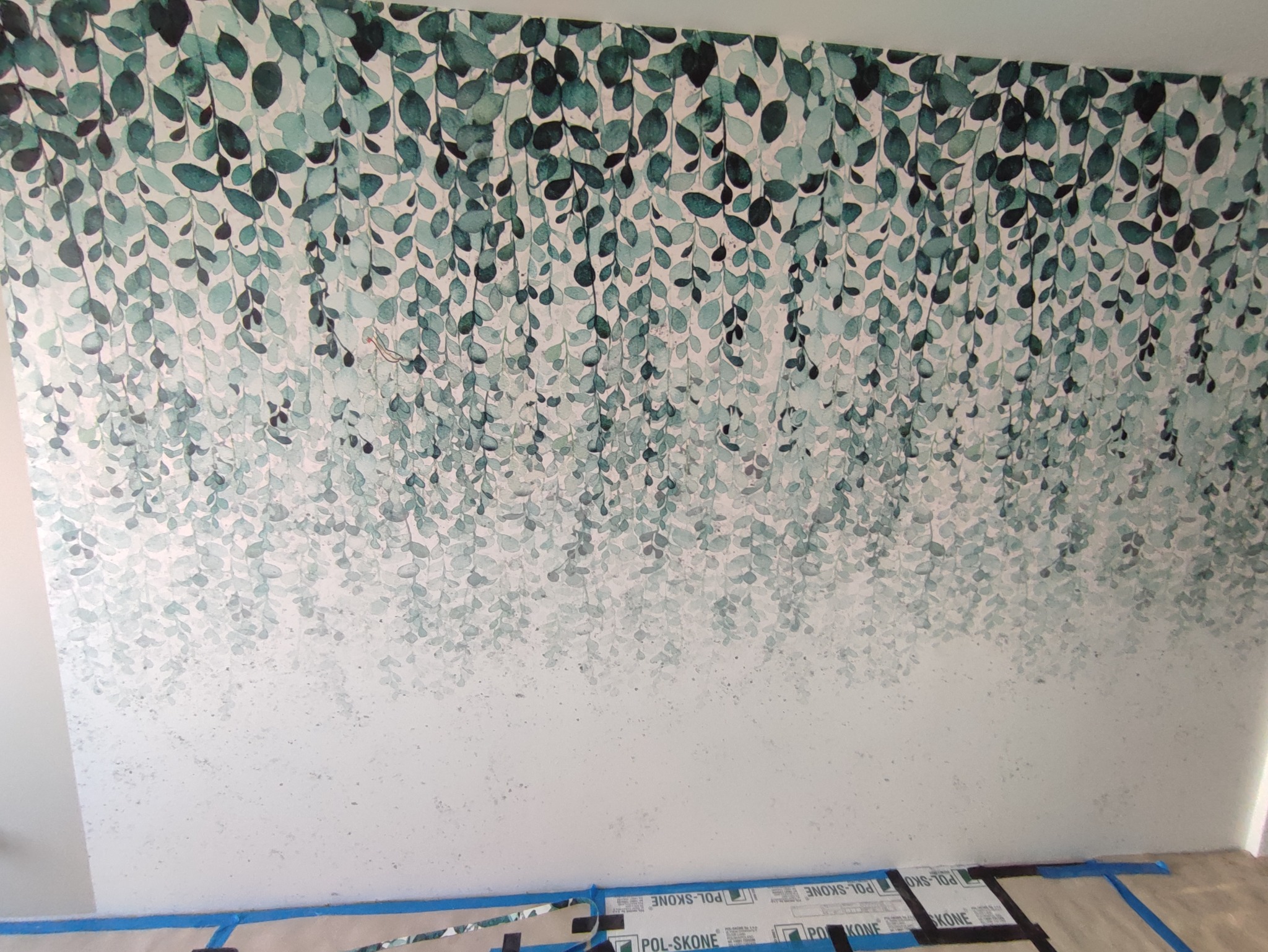 Ściana z tapetą w motywie zwisających, akwarelowych liści w odcieniach zieleni i bieli, zabezpieczona na dole taśmą malarską i kartonami ochronnymi.