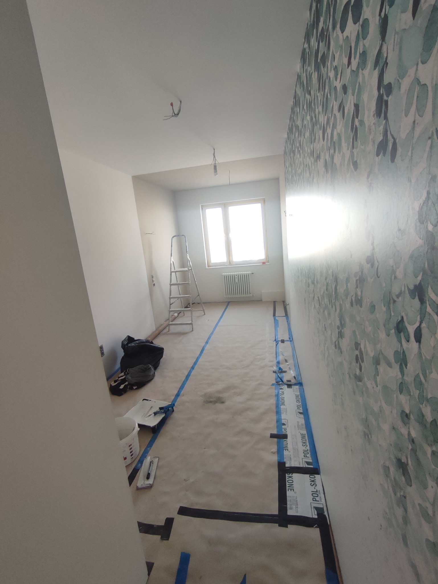 Pokój w trakcie odświeżania: ściana z tapetą w liście, podłoga zabezpieczona folią malarską i taśmą, drabina oparta o ścianę, widoczne narzędzia malarskie i wiadro z farbą.