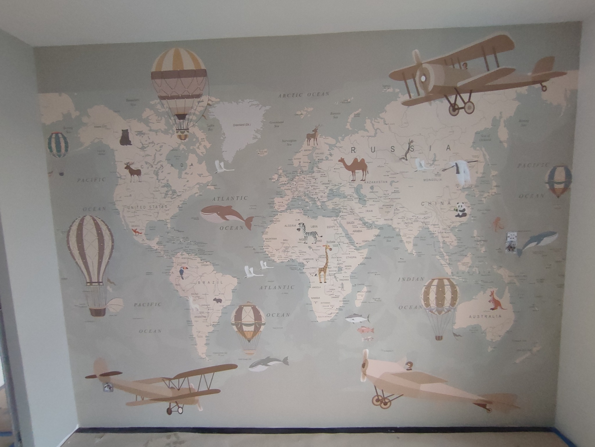 Dziecięca tapeta z mapą świata, na której widnieją ilustracje zwierząt, balonów i samolotów w pastelowych odcieniach beżu i szarości.