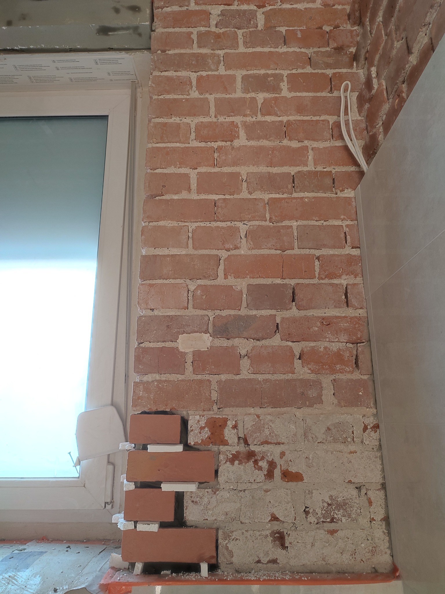 Fragment ściany z czerwonej cegły w trakcie renowacji, widoczne świeżo ułożone cegły z użyciem białych podkładek dystansowych, częściowo otynkowana dolna część ściany, okno z widocznym profilem...