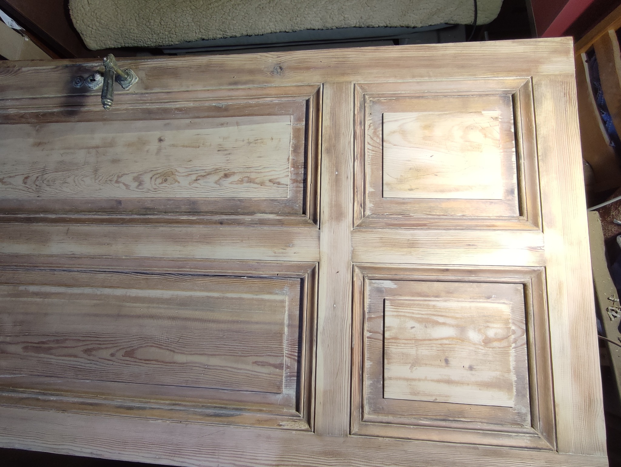 Drewniane drzwi z czterema panelami w trakcie renowacji, z widocznymi śladami starej farby i nowymi warstwami bejcy, z zamontowaną starą klamką.