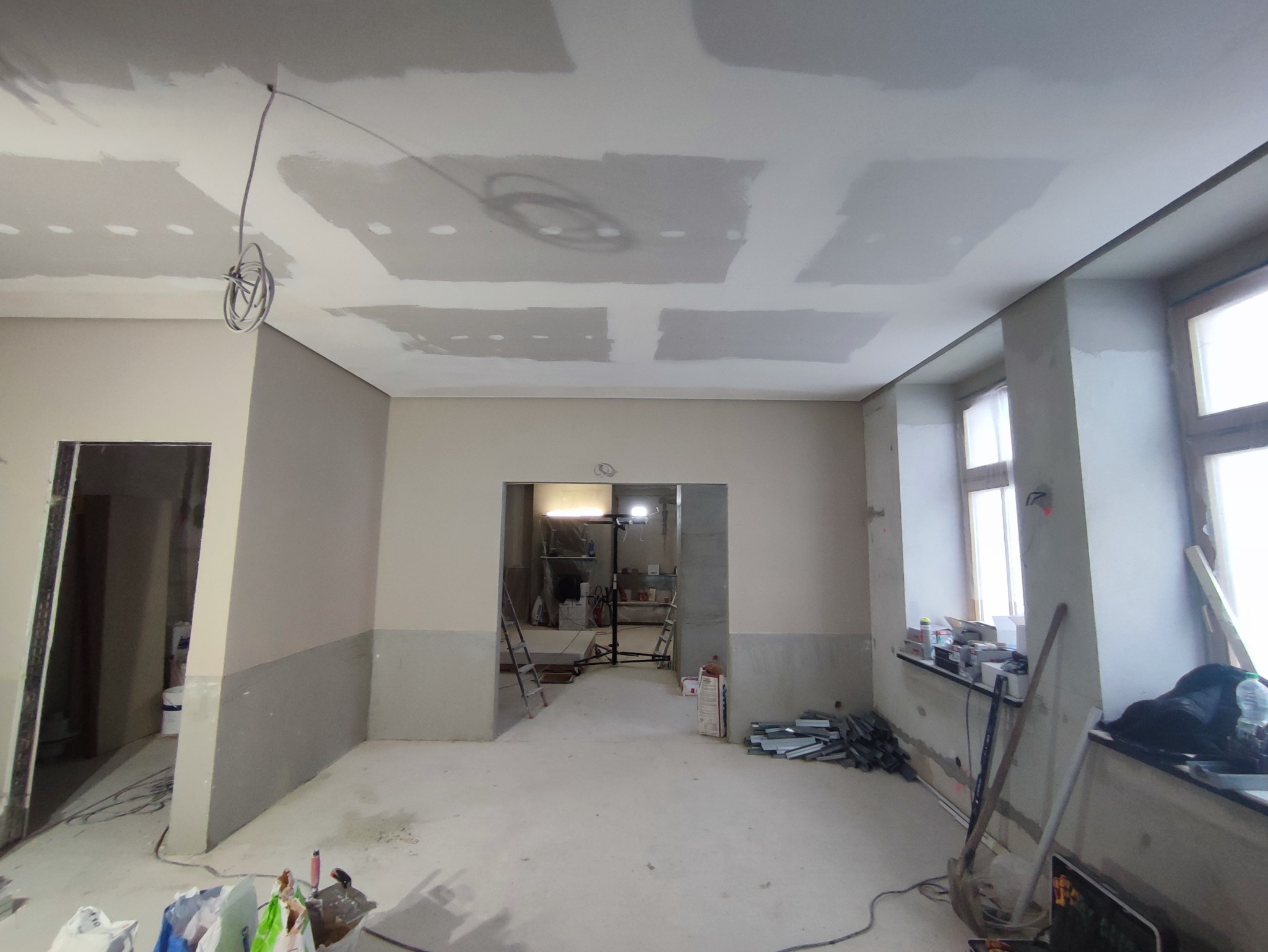 Wnętrze w trakcie prac wykończeniowych, widoczne płyty gipsowo-kartonowe na suficie, szpachlowane ściany do połowy wysokości, przewody elektryczne zwisające z sufitu, oraz narzędzia i materiały...