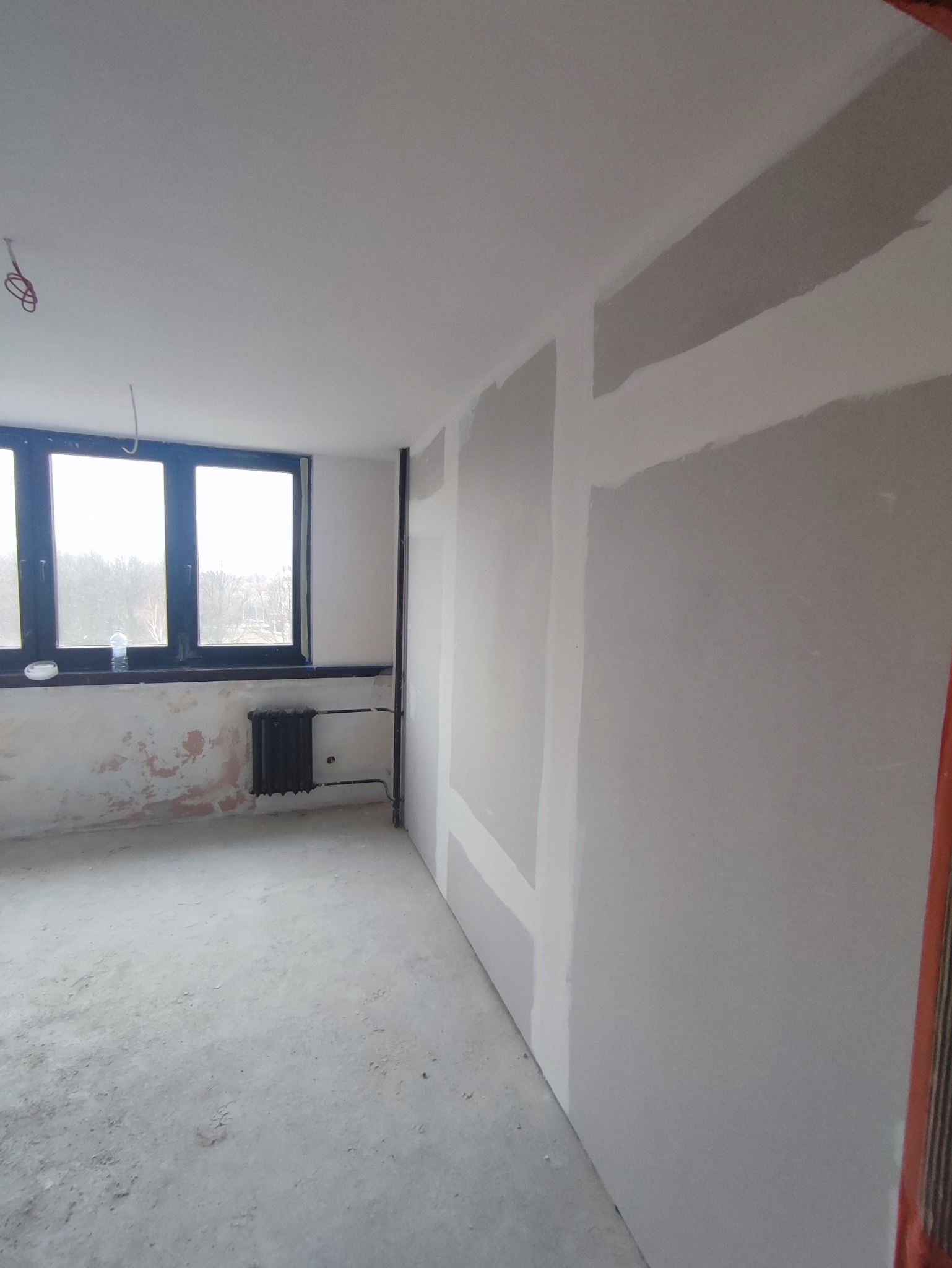 Wnętrze pokoju we Wrocławiu w trakcie prac remontowych, widoczne płyty gipsowo-kartonowe na ścianach, przygotowane do szpachlowania i malowania, oraz stary grzejnik pod oknem.