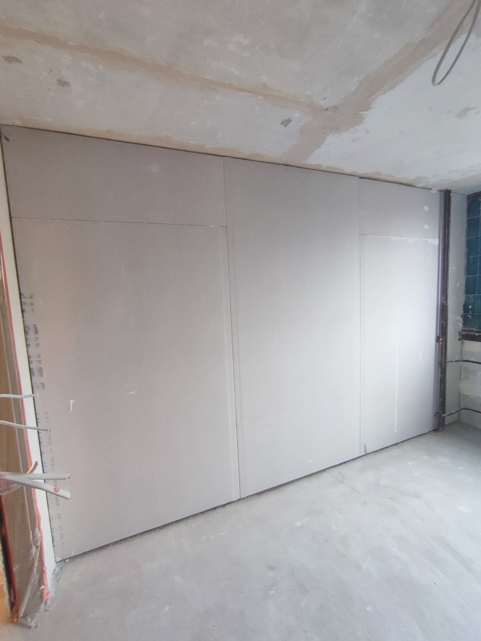 Ściana z płyt gipsowo-kartonowych w trakcie montażu, widoczne łączenia płyt, surowy betonowy sufit i podłoga, instalacja elektryczna przy ścianie.