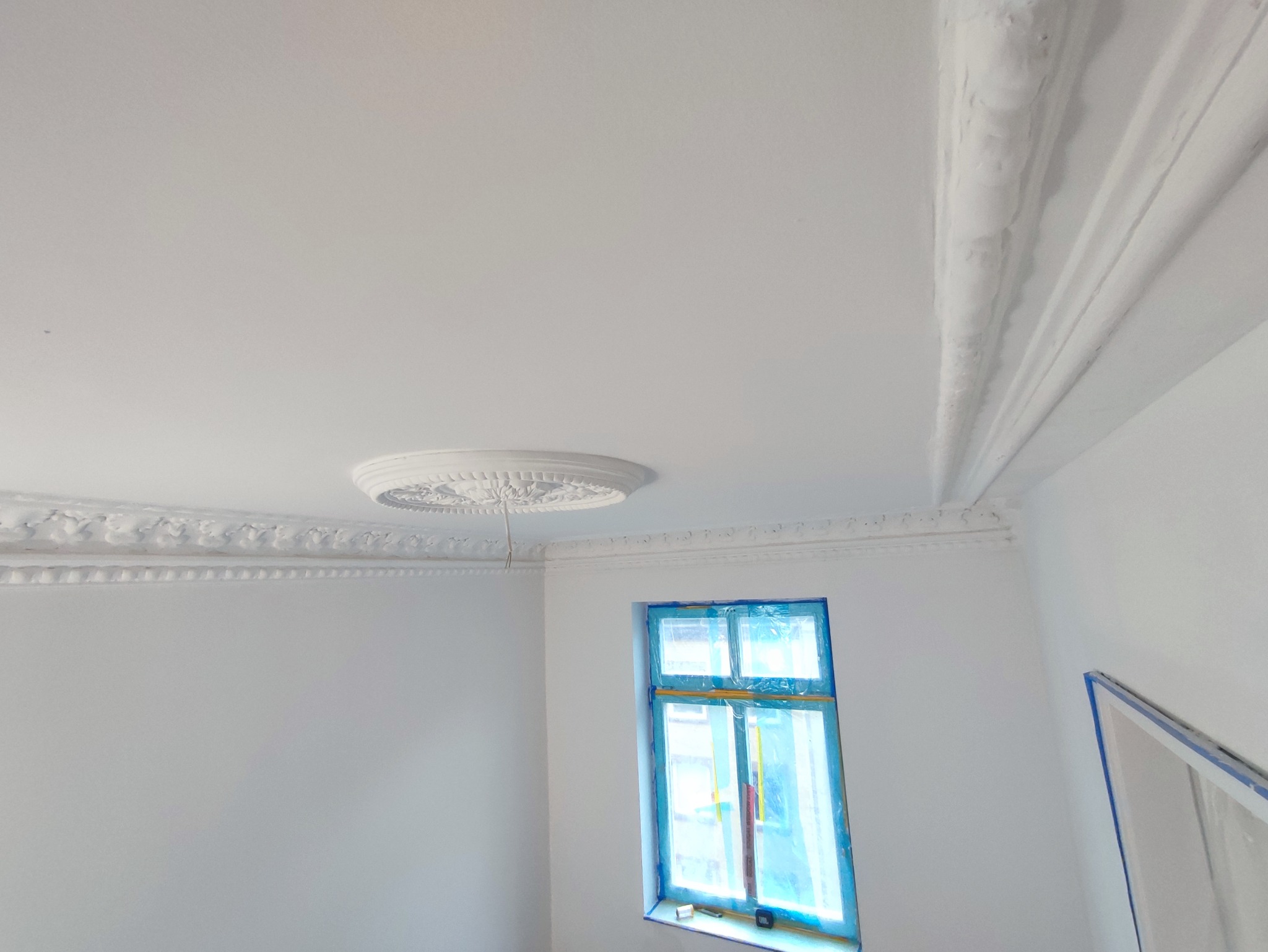 Białe ściany i sufit z dekoracyjnymi listwami sztukateryjnymi i rozetą sufitową, okno zabezpieczone folią malarską.
