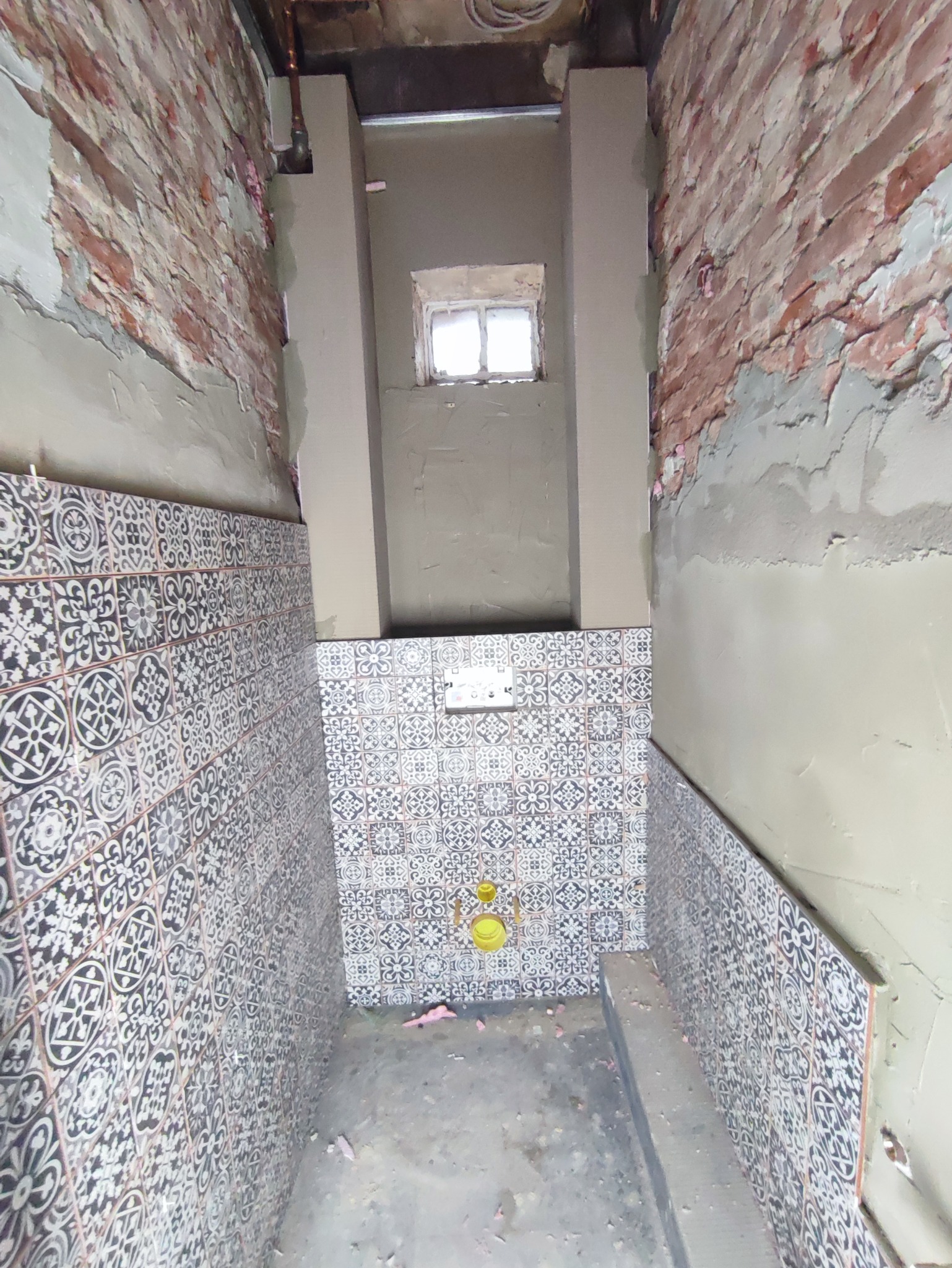 Wnętrze łazienki w trakcie prac, widoczne nowe płytki z wzorem na ścianach, surowa cegła na górze ścian, pionowe elementy konstrukcyjne obudowane płytami oraz małe okno w głębi.