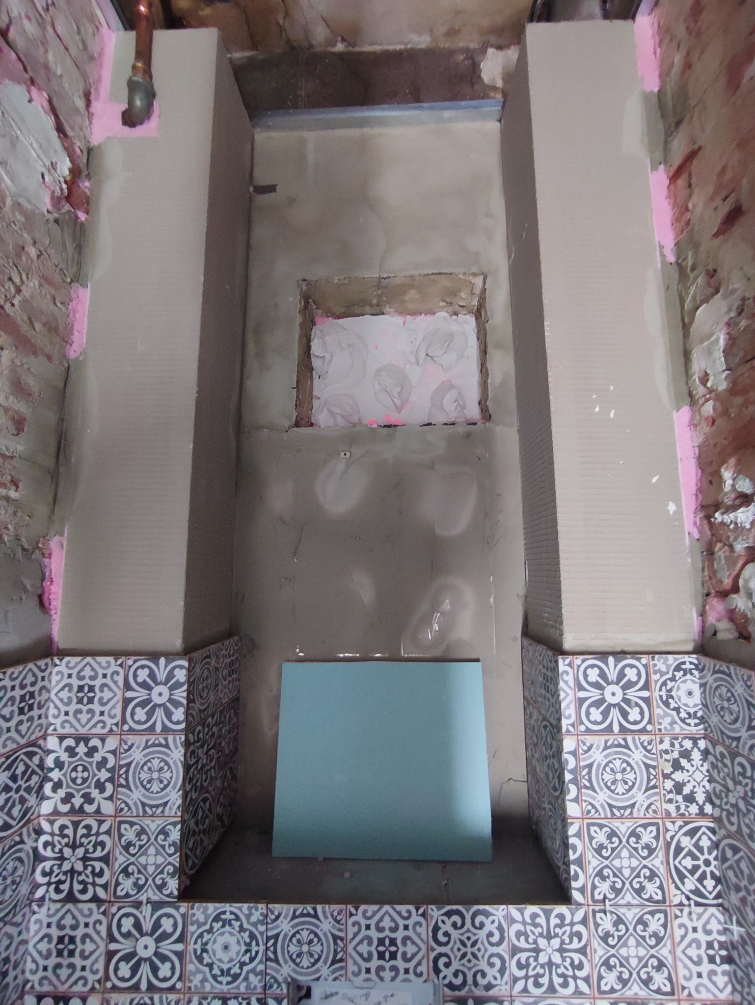 Wnętrze łazienki w trakcie prac: widoczne fragmenty surowej ściany, pionowe profile obłożone płytkami w stylu retro, zamurowane okienko z resztkami tapety, oraz niebieska płyta gipsowa oparta...